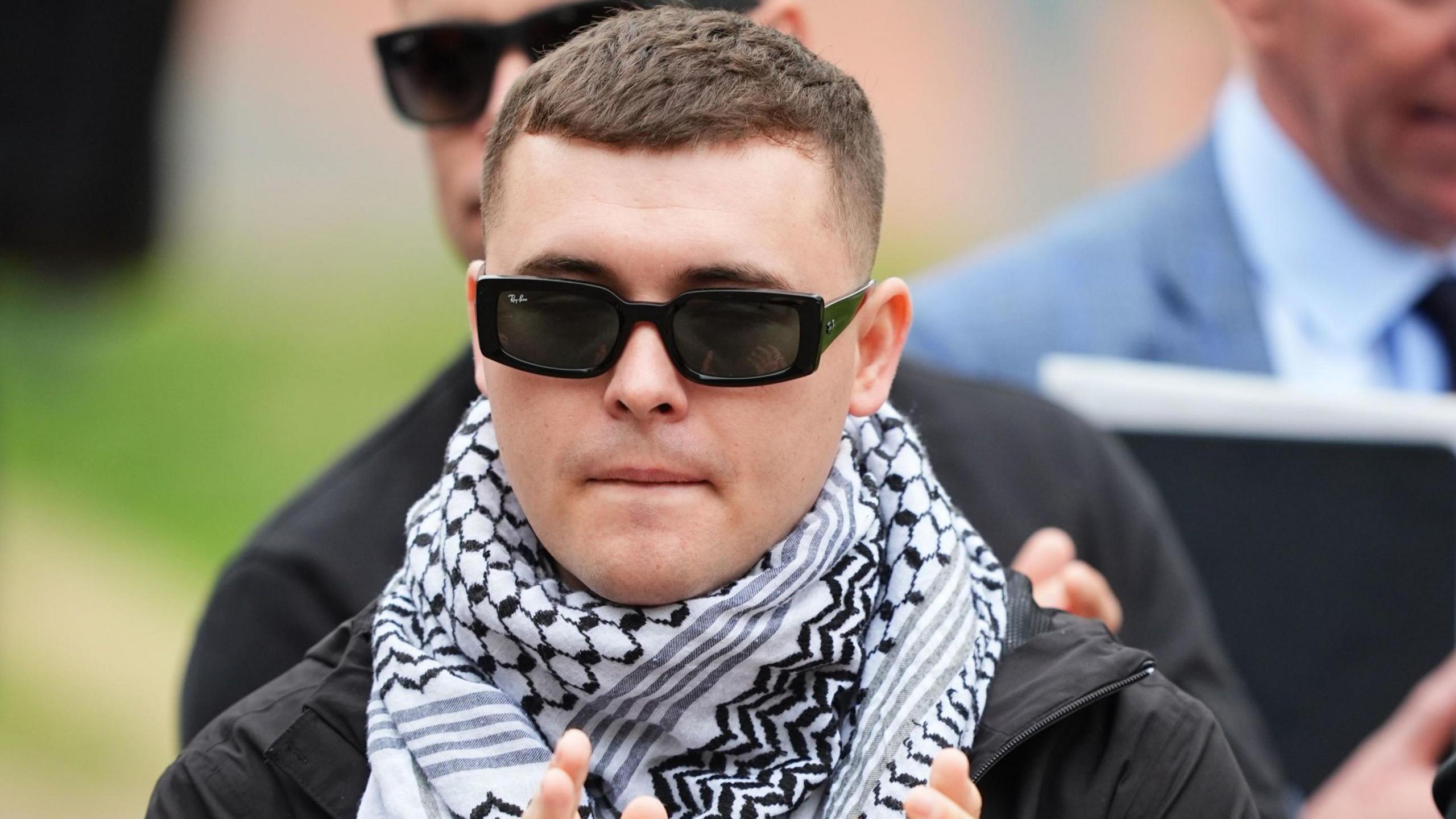 Kneecap's Liam Óg Ó hAnnaidh, who performs under the stage name Mo Chara, outside Woolwich Crown Court, London. He is wearing black sunglasses and a white and black keffiyeh. He has dark hair.