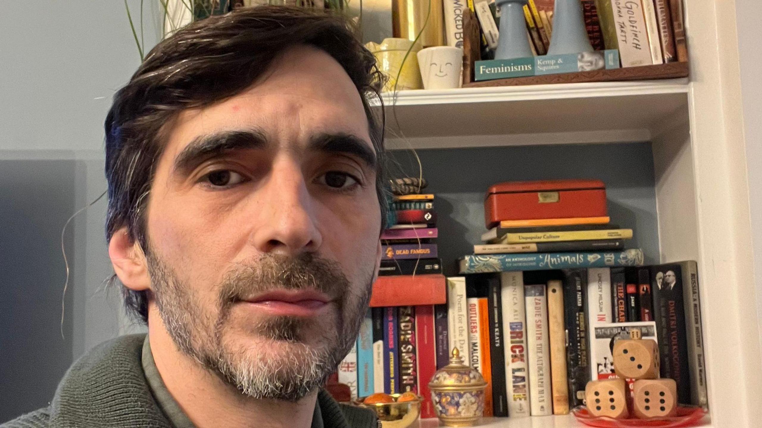 Omar is standing in front of a bookcase. He is looking solemnly into the camera. 
