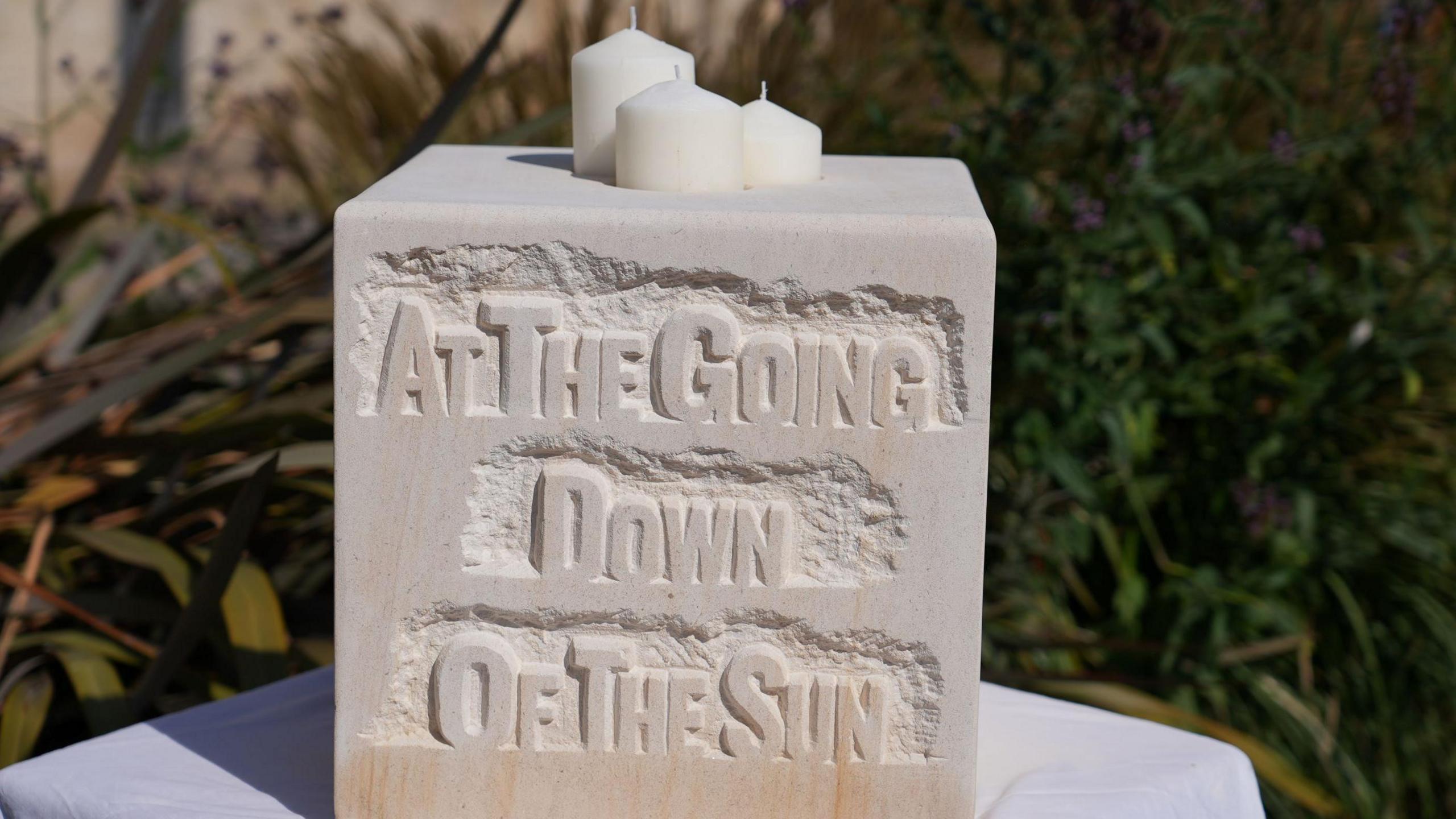 A stone carving stands on a table in front of foliage. It reads "at the going down of the sun" and has a hole where three white candles have been inserted at the top.