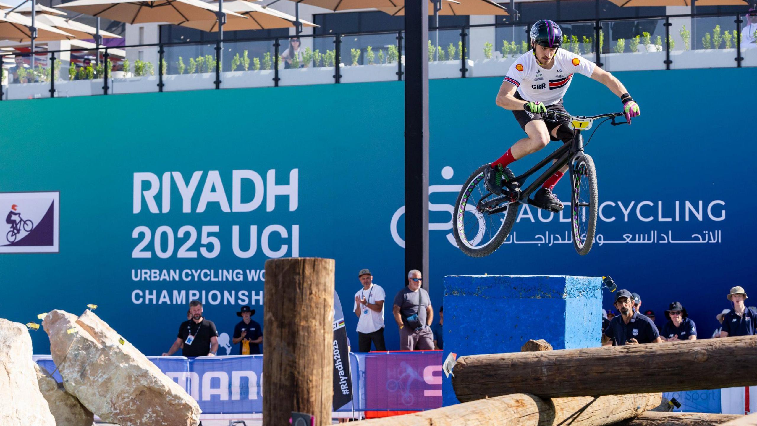 Rolls jumps his bike over a large and tall blue box on a bike trial course. He wears a blue helmet, white top, black shorts, red socks and light coloured gloves. Spectators watch on from the side of the course. A large blue board surrounds the course with the name of the event printed on it. 