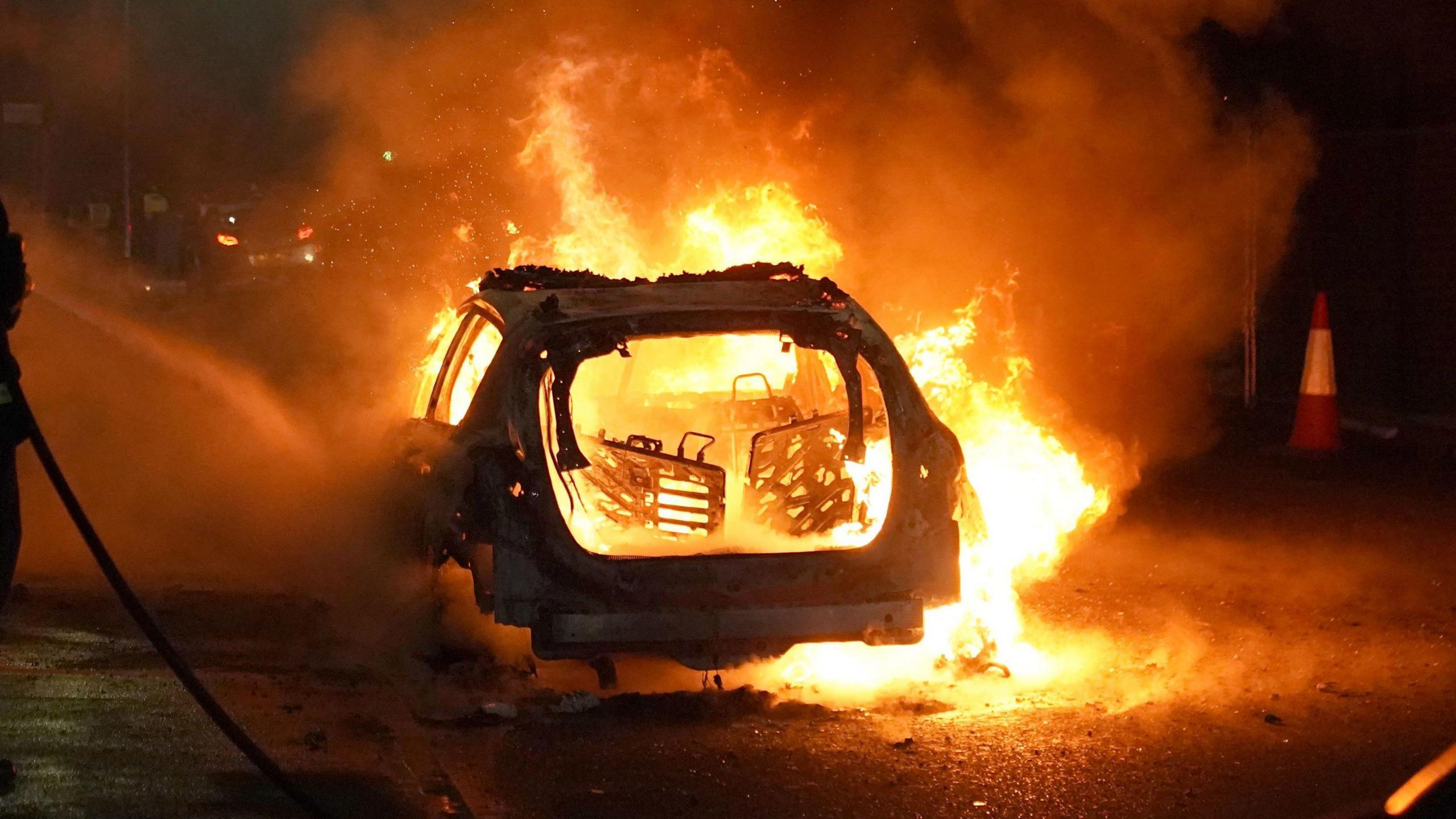 Burning car in the night. There is fire all around and the car is charred with the back completely gone and fire engulfing the inside and the seats.