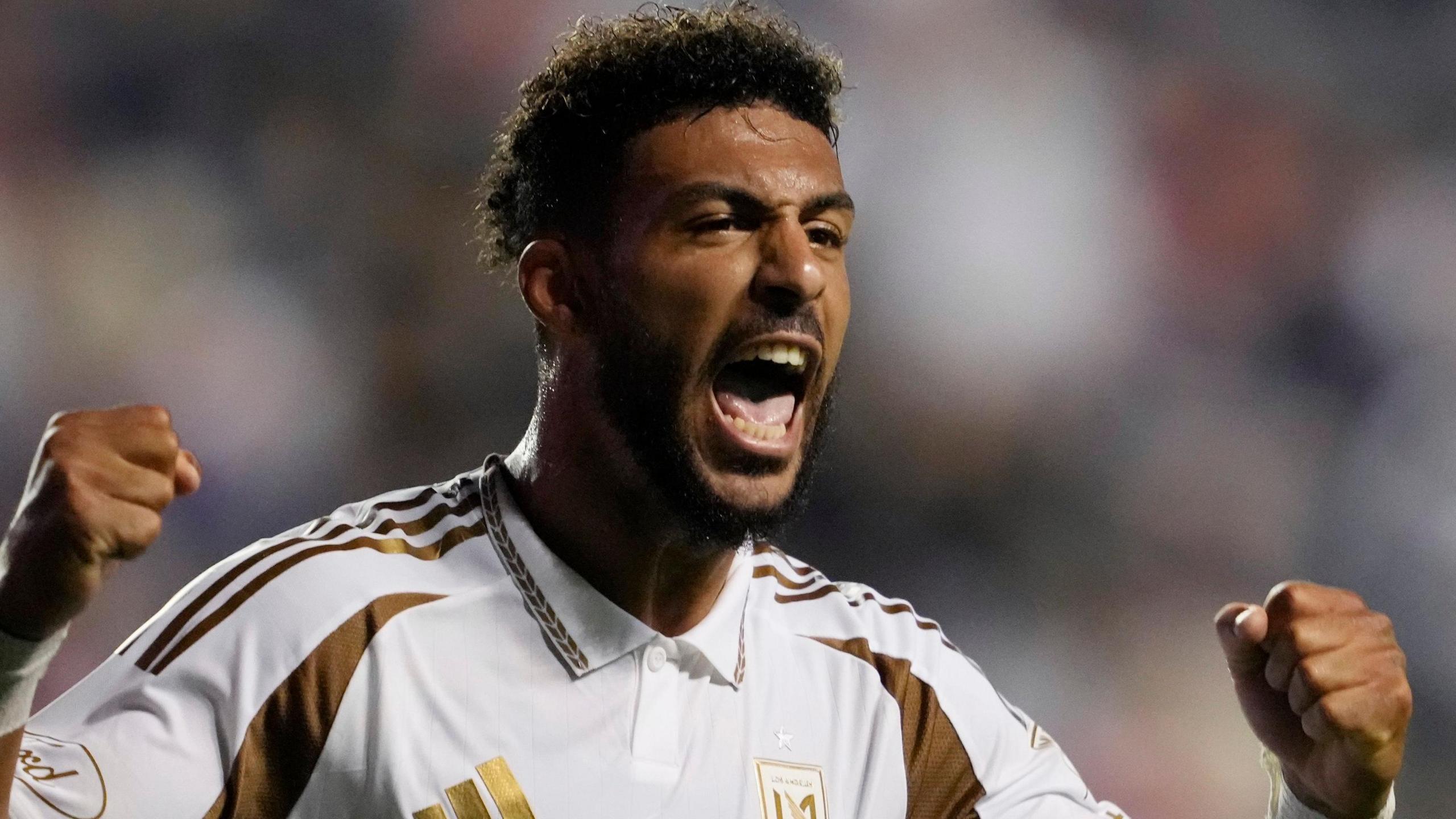 Denis Bouanga, wearing a white and gold LA FC shirt, holds both fists up in triumph and shouts as he celebrates a goal