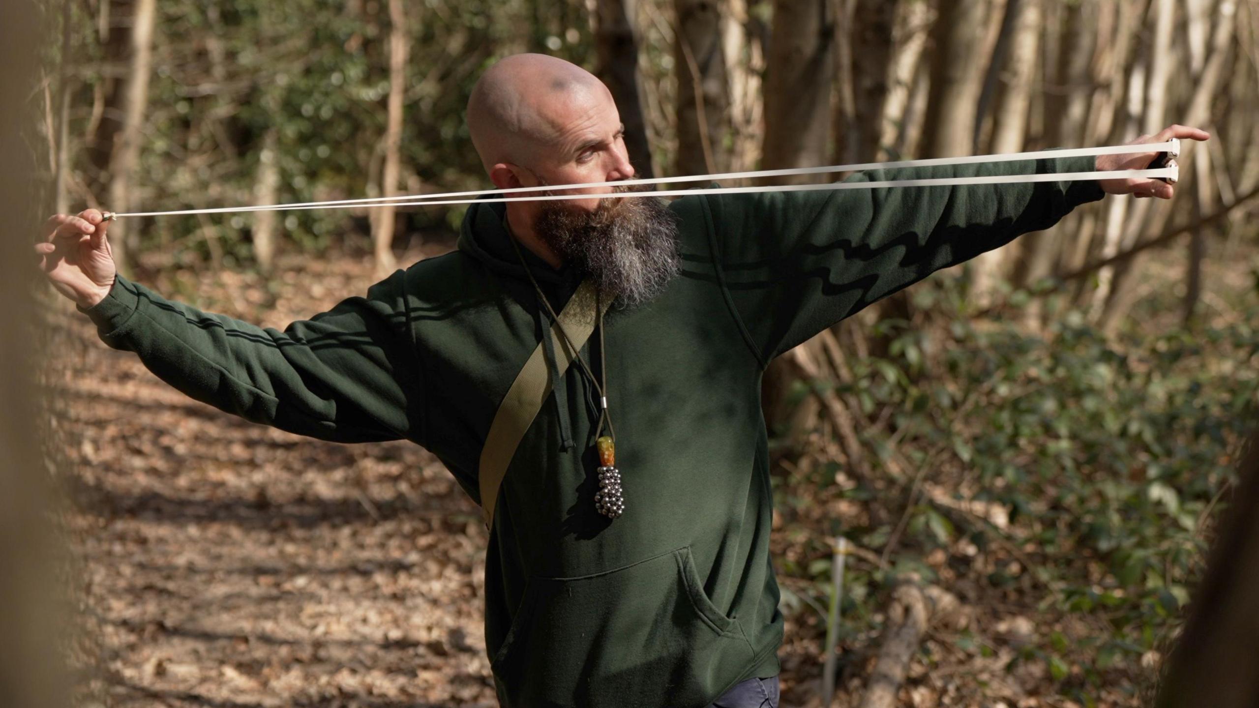 A side profile photo of a man in a wooded setting aiming a catapult with the elastic pulled back.
