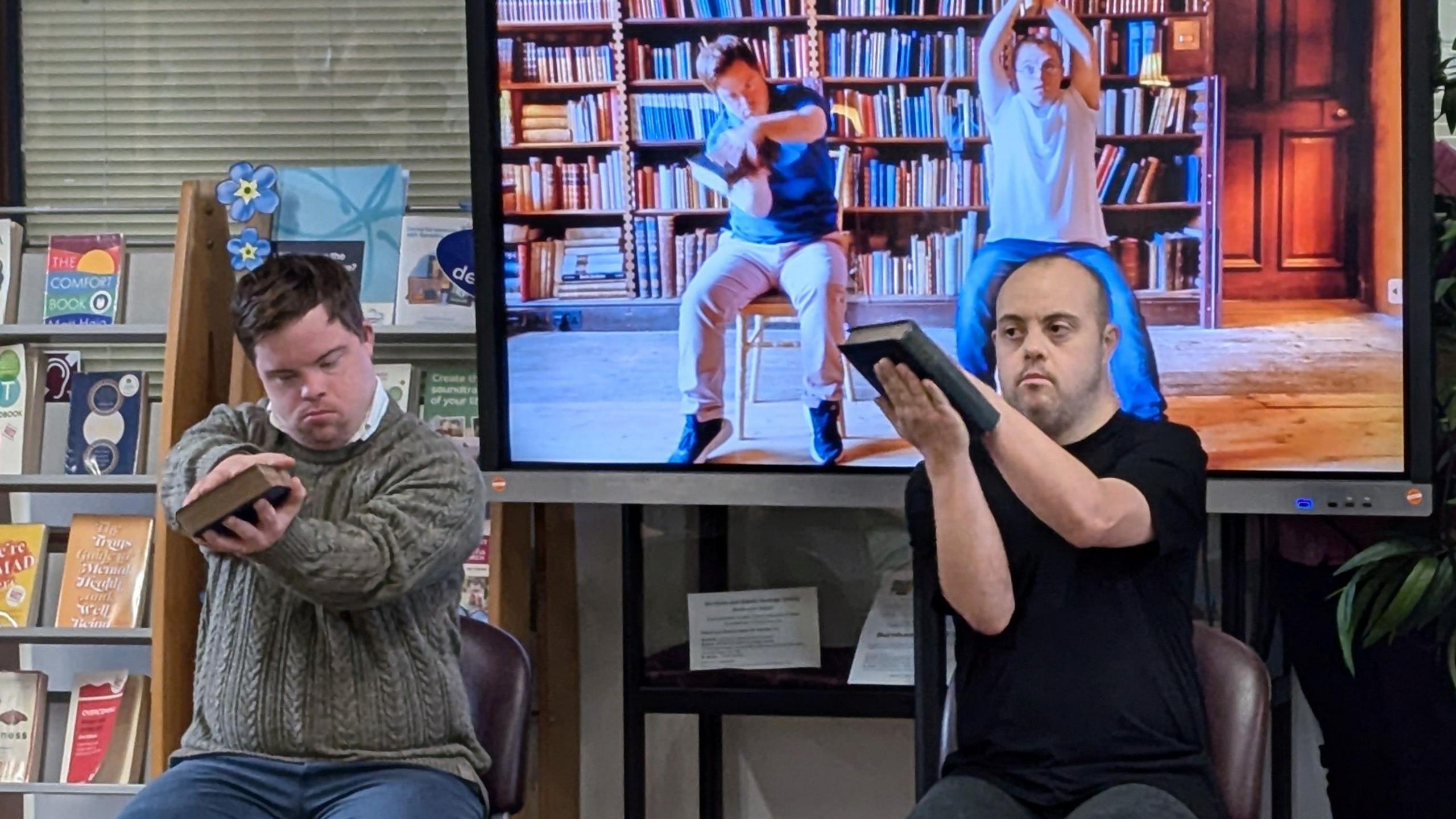 A picture of two men, who are sitting down on chairs in front of a TV screen which also has two men on screen sitting on chairs. One man has a grey-green jumper on with blue trousers; the other a black jumper and jeans.