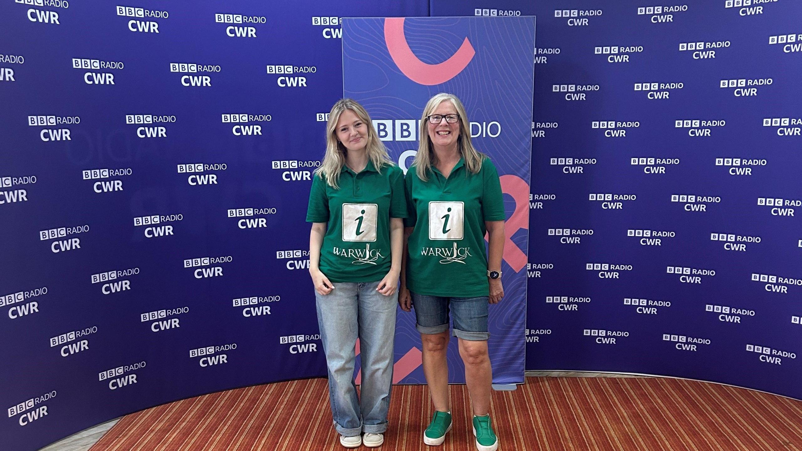 Two women stand in front of purple BBC Coventry and Warwickshire branding. They are both blonde and are wearing green tshirts. There is a gold logo on them with the letter "i". Below the logo, it reads "Warwick"