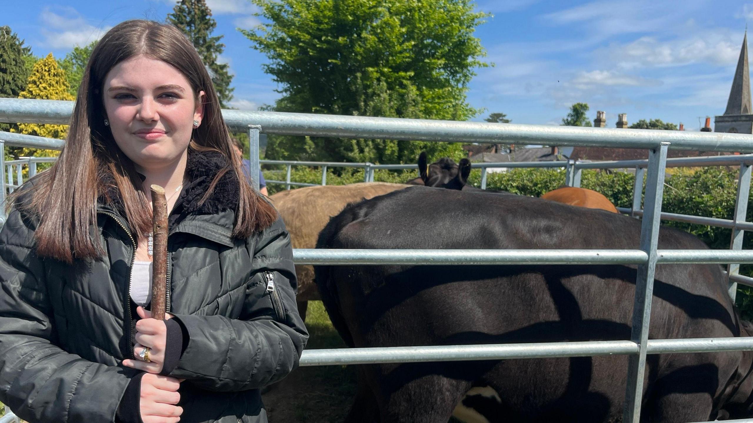 A young woman with shoulder length dark hair and and wearing a black jacket is standing in front of a pen with cows inside. None of them are looking at the camera. A church spire can be seen in the background.