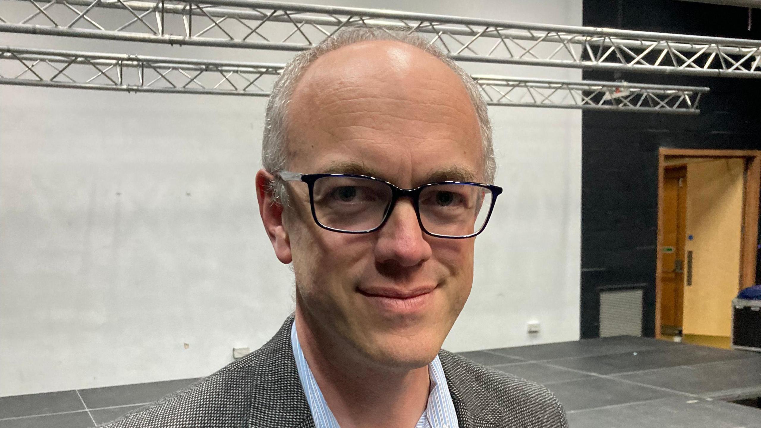 Andrew Lister from the shoulders up. He is wearing a tweed-looking jacket and shirt, as well as glasses. Behind him is a stage, metal bars and a white wall.