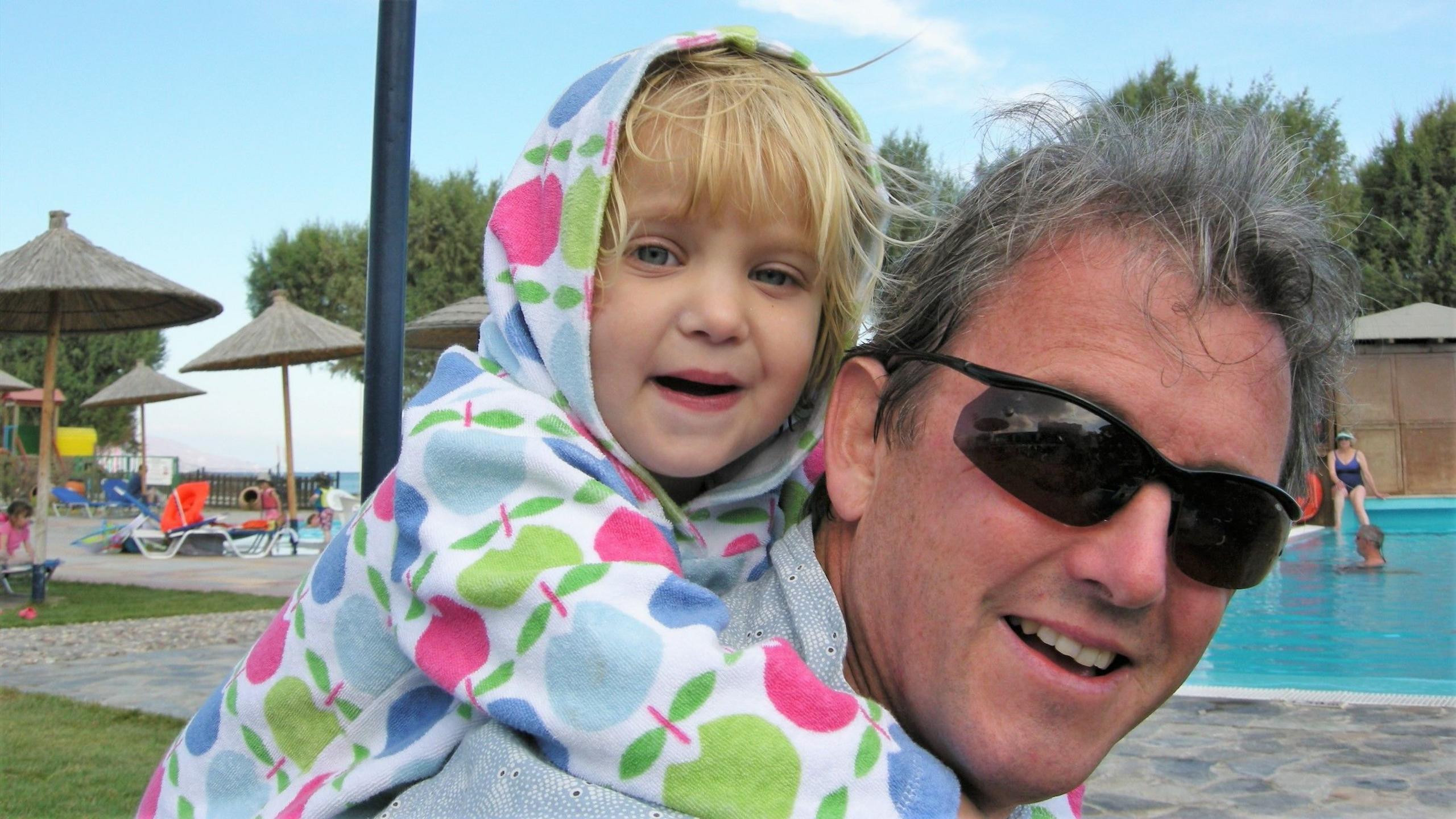 Mike Calnan holding his infant daughter Flossy, who is wearing a colourful towel onesie covered in apples. She is blonde with blue eyes and is hugging her dad over his shoulders, who is wearing dark sunglasses and has grey hair. They are beside a pool with lots of sun loungers and straw parasols. 