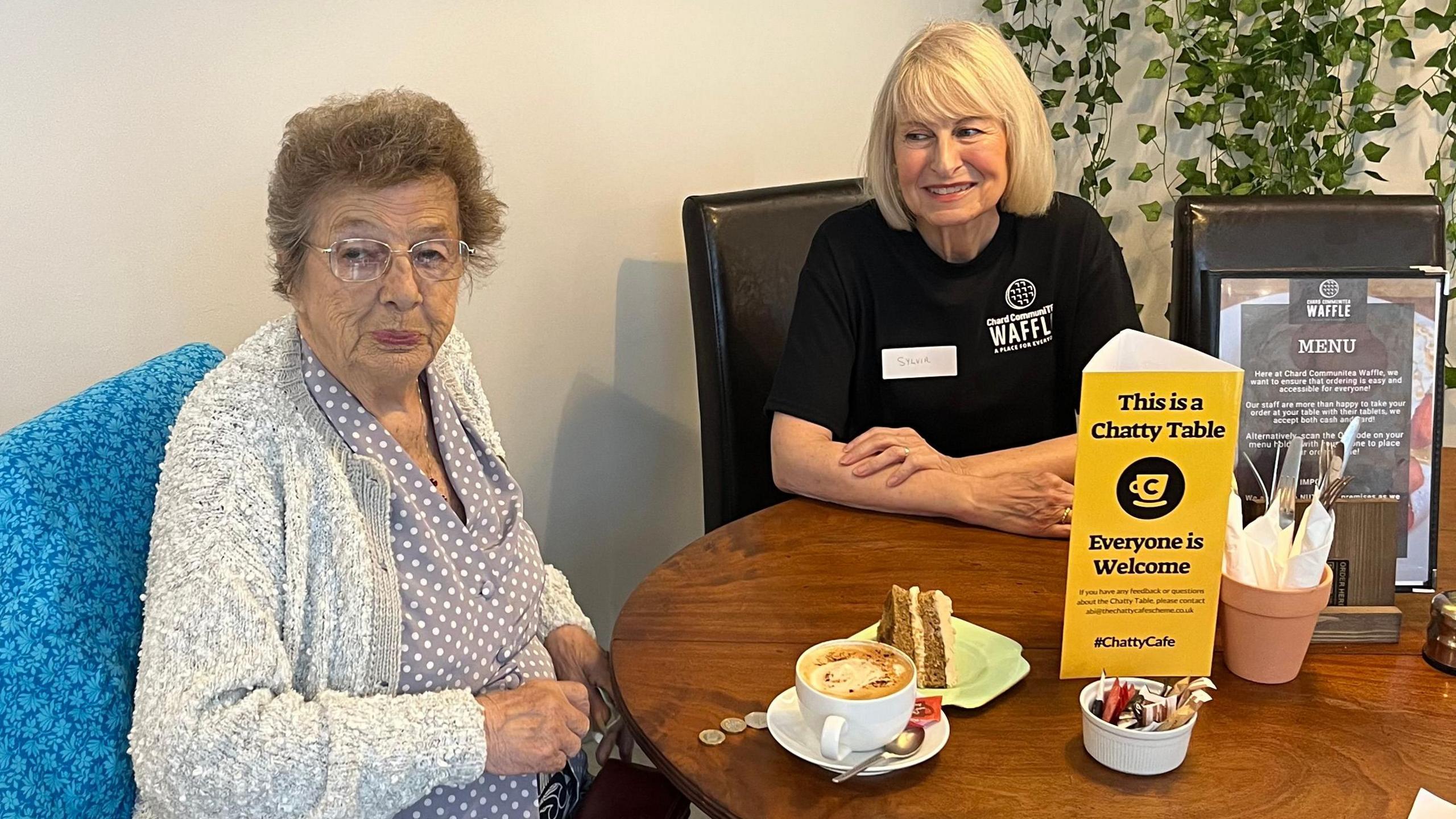 Two ladies sit at a table. An older lady who's wearing glasses is dressed in a blue shirt with white polkadots and a white and grey cardigan. In front of her there's a slide of cake and a coffee. Next to her there's a slightly younger lady wearing a volunteer shirt who smiles adoringly at her. On the table there is a triangular sign that says 'This is a Chatty Table".