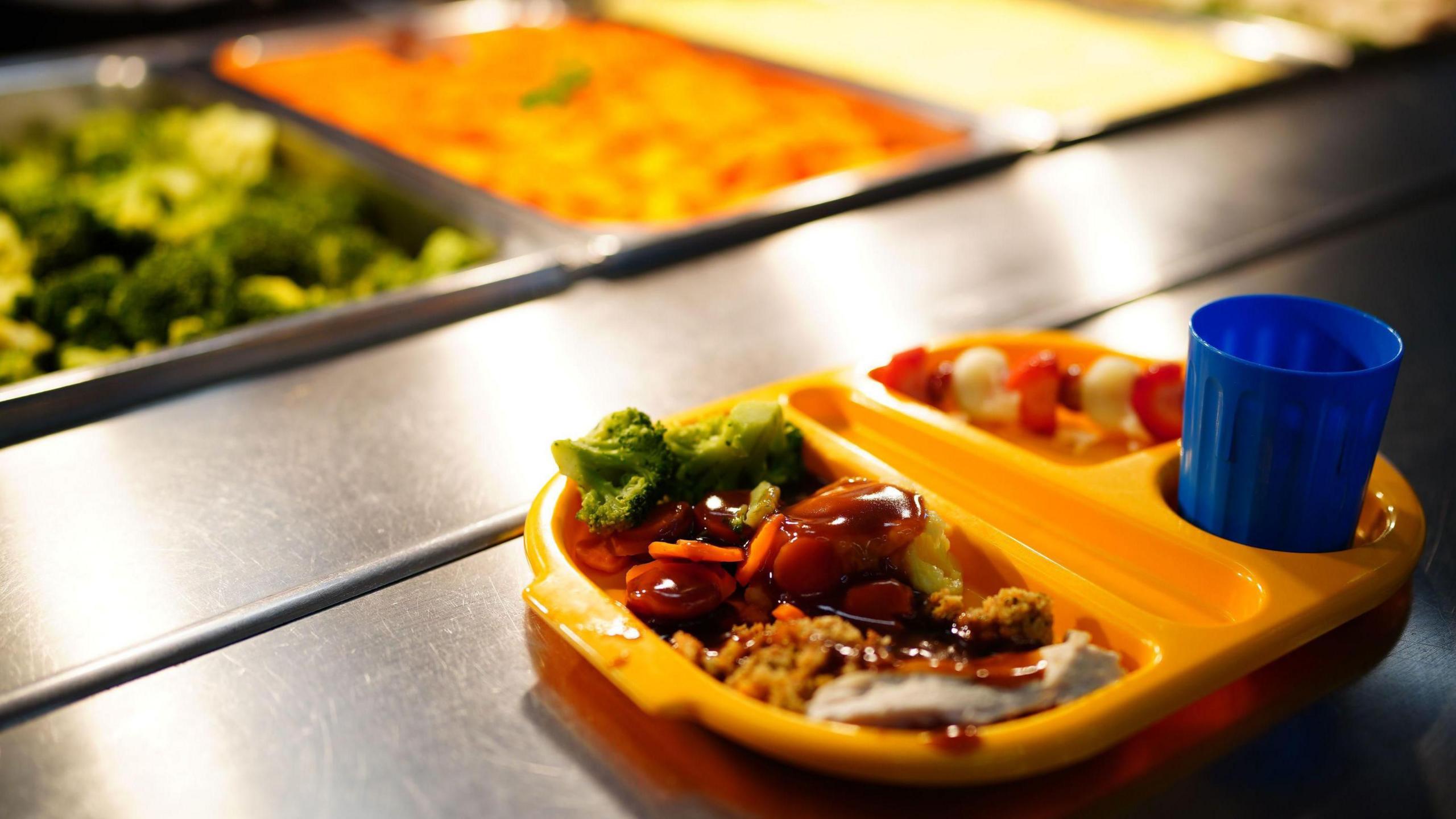 A well lit school dinner in a round, yellow plastic tray in front of a hot plate of broccoli. It has sausage, broccoli, mash and gravy. 