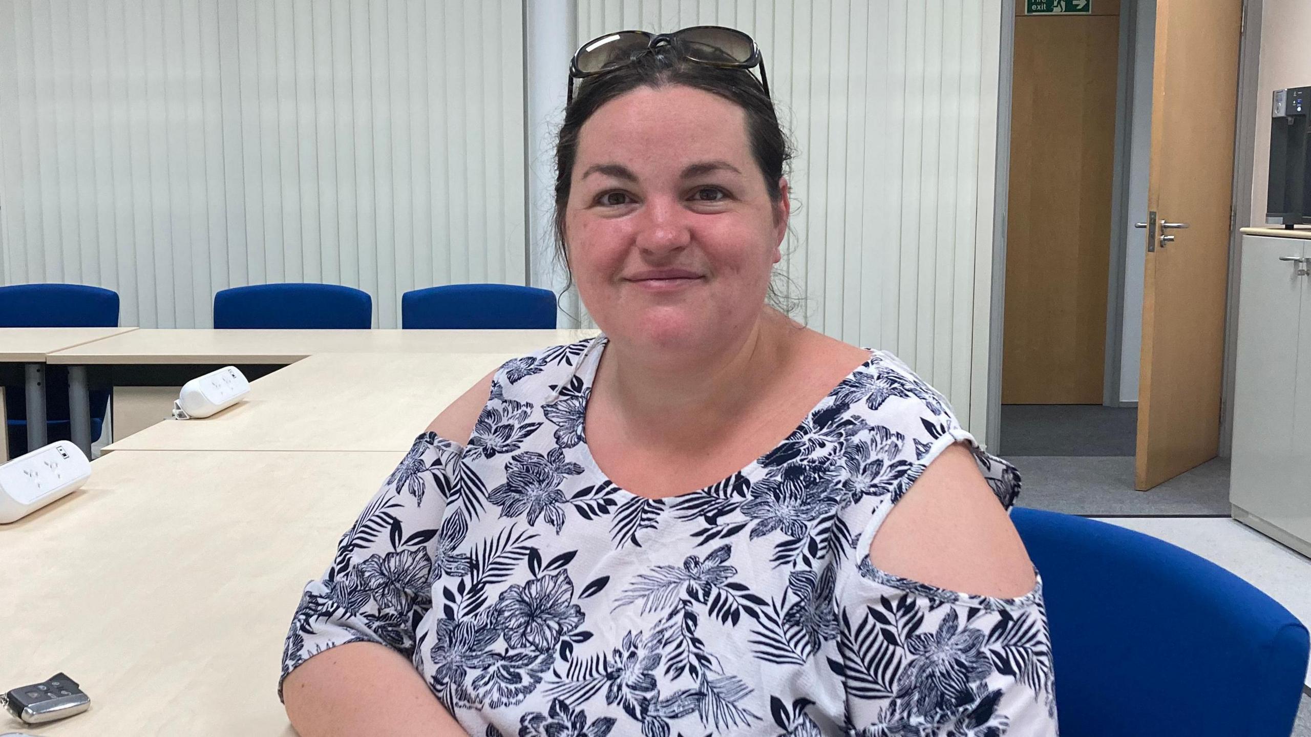 Heather Williams smiles at the camera. She wears a white top with a navy flowery pattern on it and sunglasses on her head. She sits on a blue office chair next to a beige table.