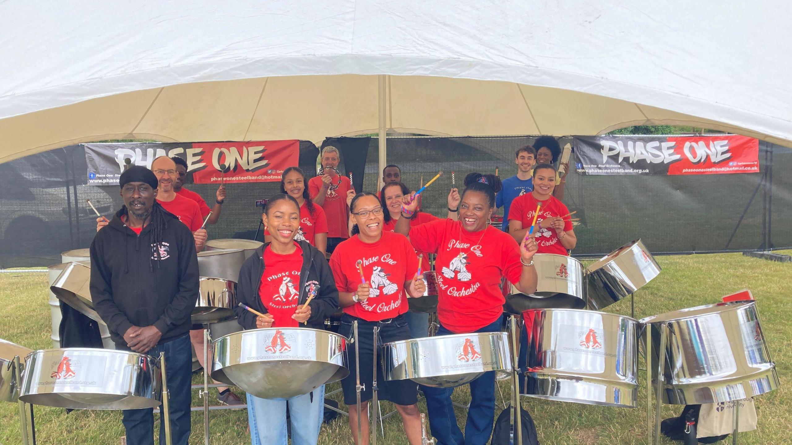 A steel pan band stands with their instruments in a field. They are all wearing red t-shirts.