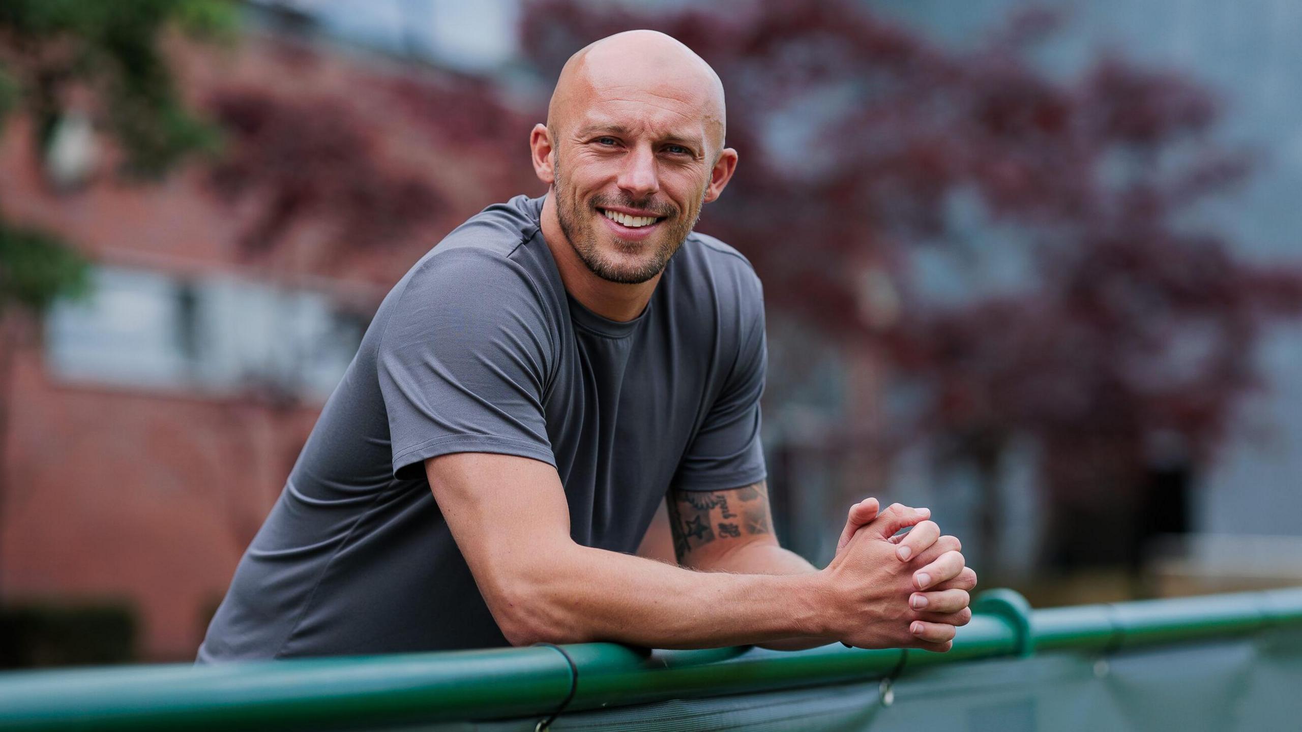 Jack Higgins leans on a metal fence. He is wearing a grey T-shirt. There is a brick building behind him in the background