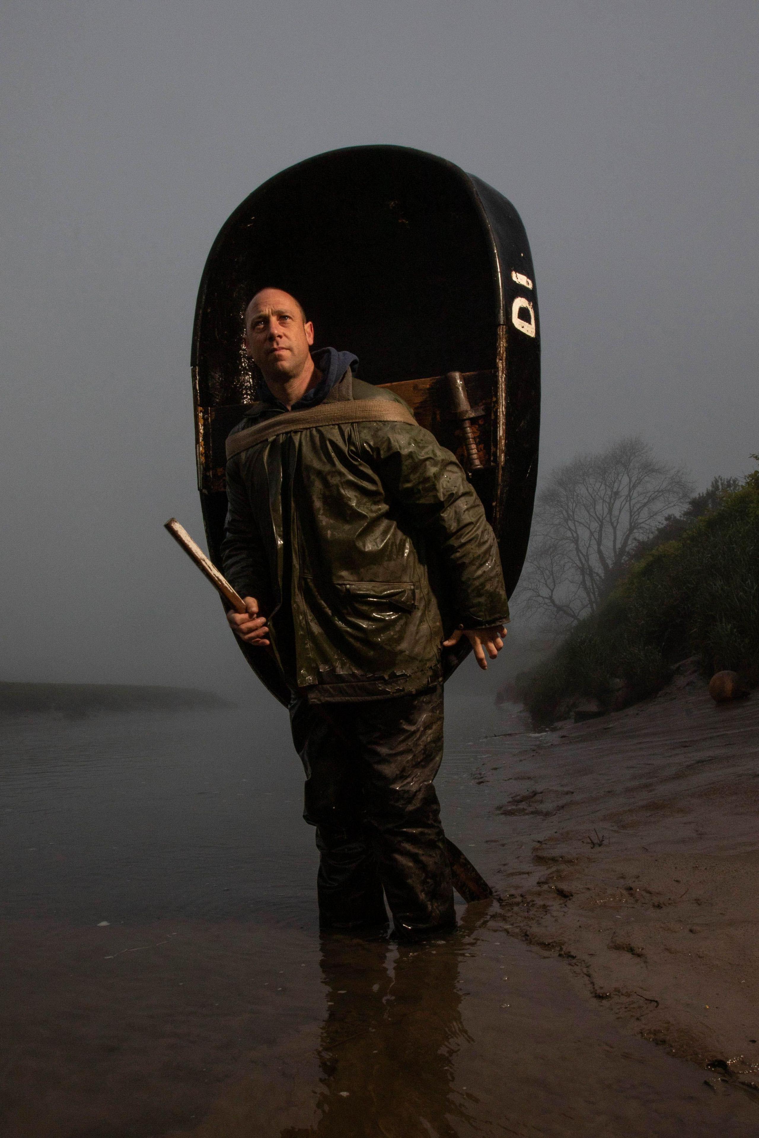 A man walking at the edge of a river bank. He has a wet clothes on and they are a dark green colour. He has a coracle on his bank and holds a rod in his hand. He has a bald head.