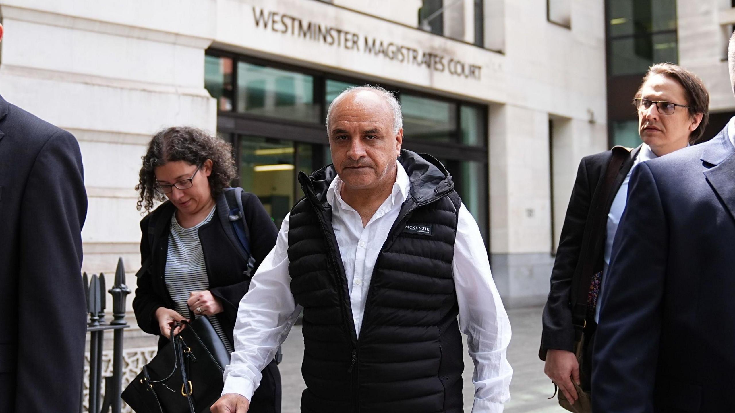 A middle aged man wearing a white shirt and a black gilet looks into the camera as he walks down the street outside a magistrates court.