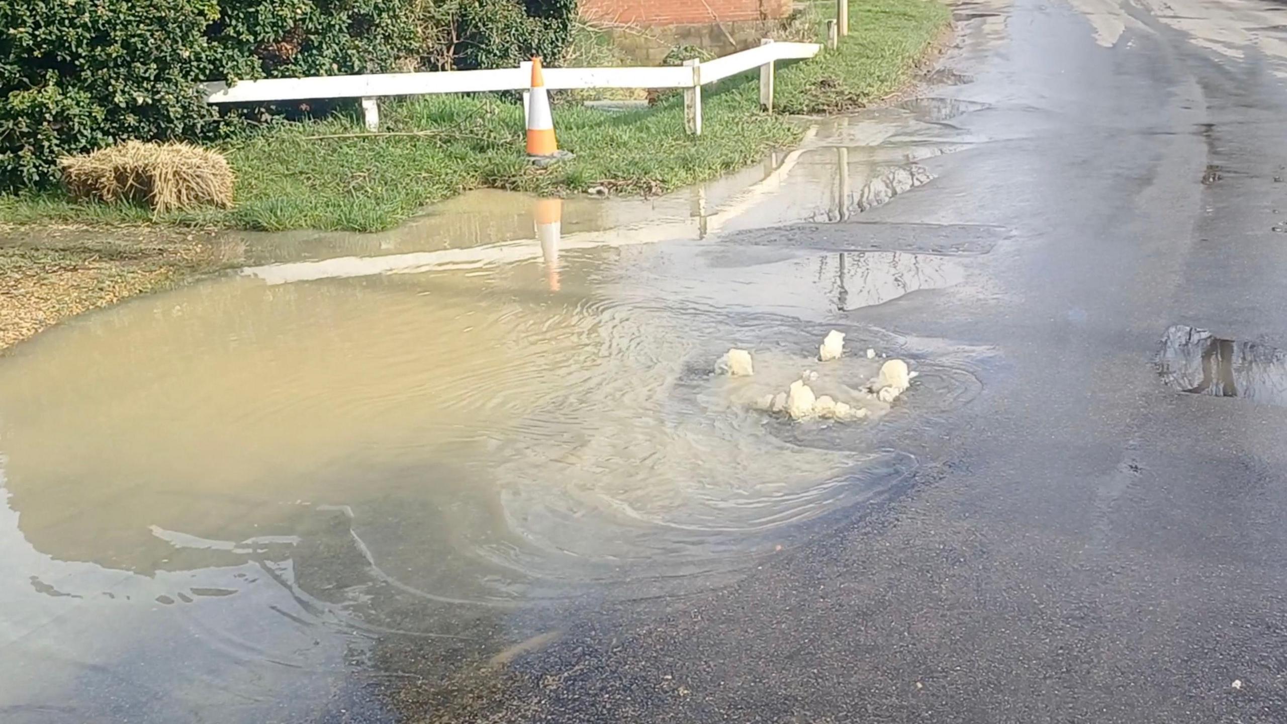 A large puddle of muddy water on a road with bubbles rising to the surface. A white fence and an orange traffic cone are on an area of grass in the background.