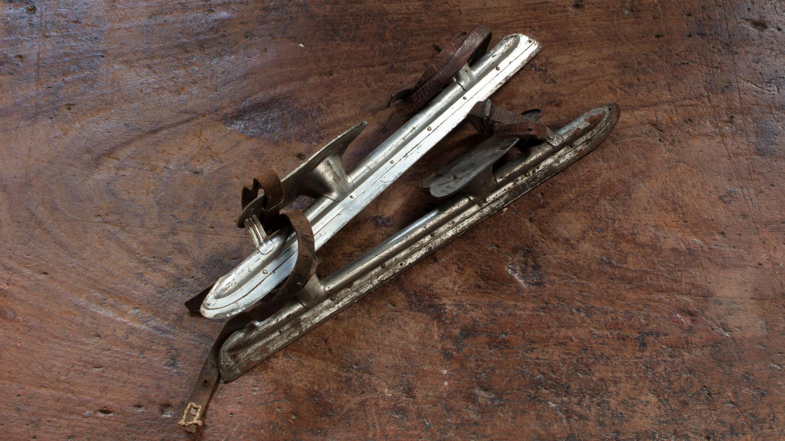 A rudimentary ice-skating blade with a platform for the foot and leather ties to strap over shoes. It is on a wooden counter.