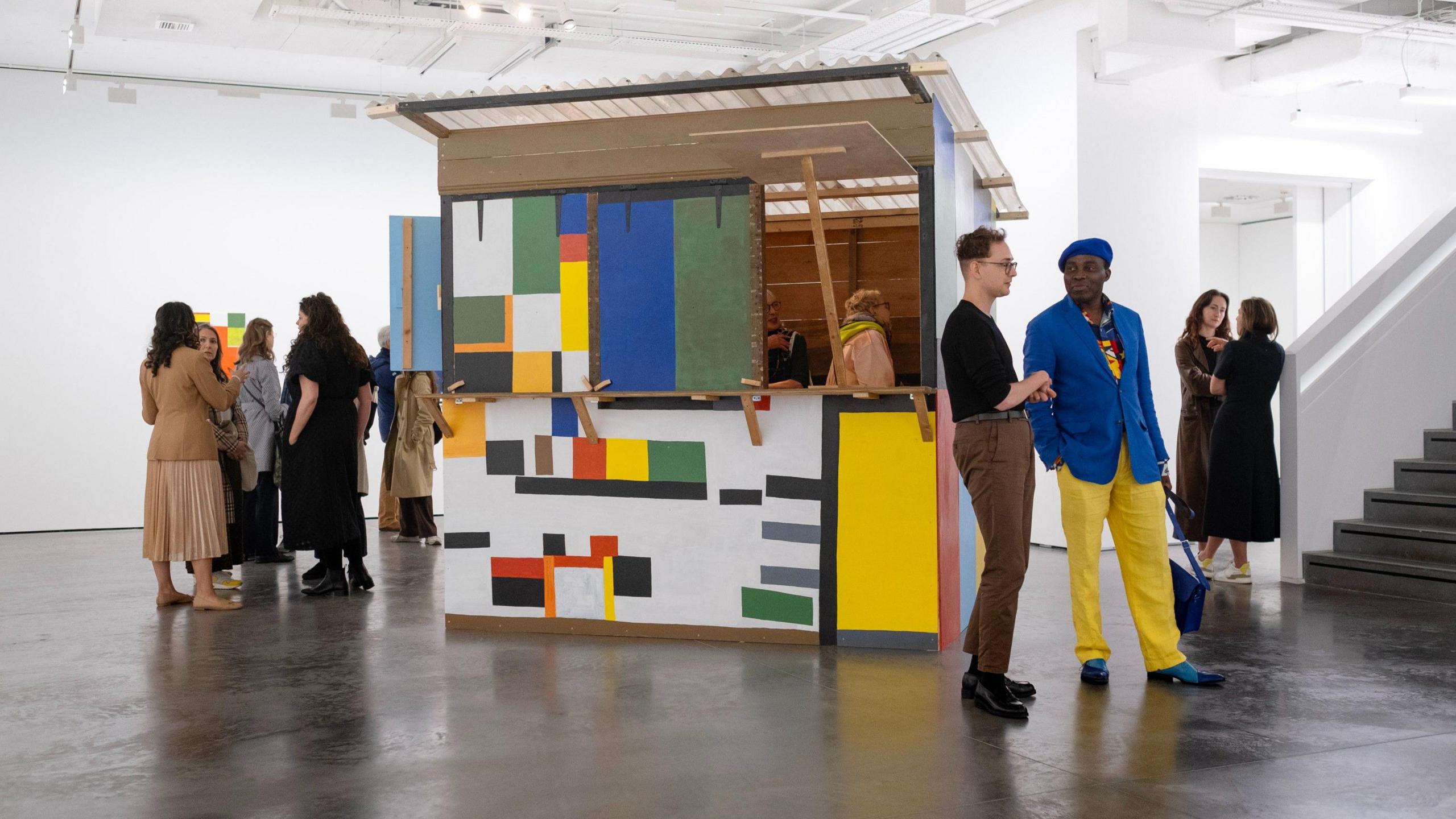 A handful of people dispersed around the gallery, around a multicoloured shack. 