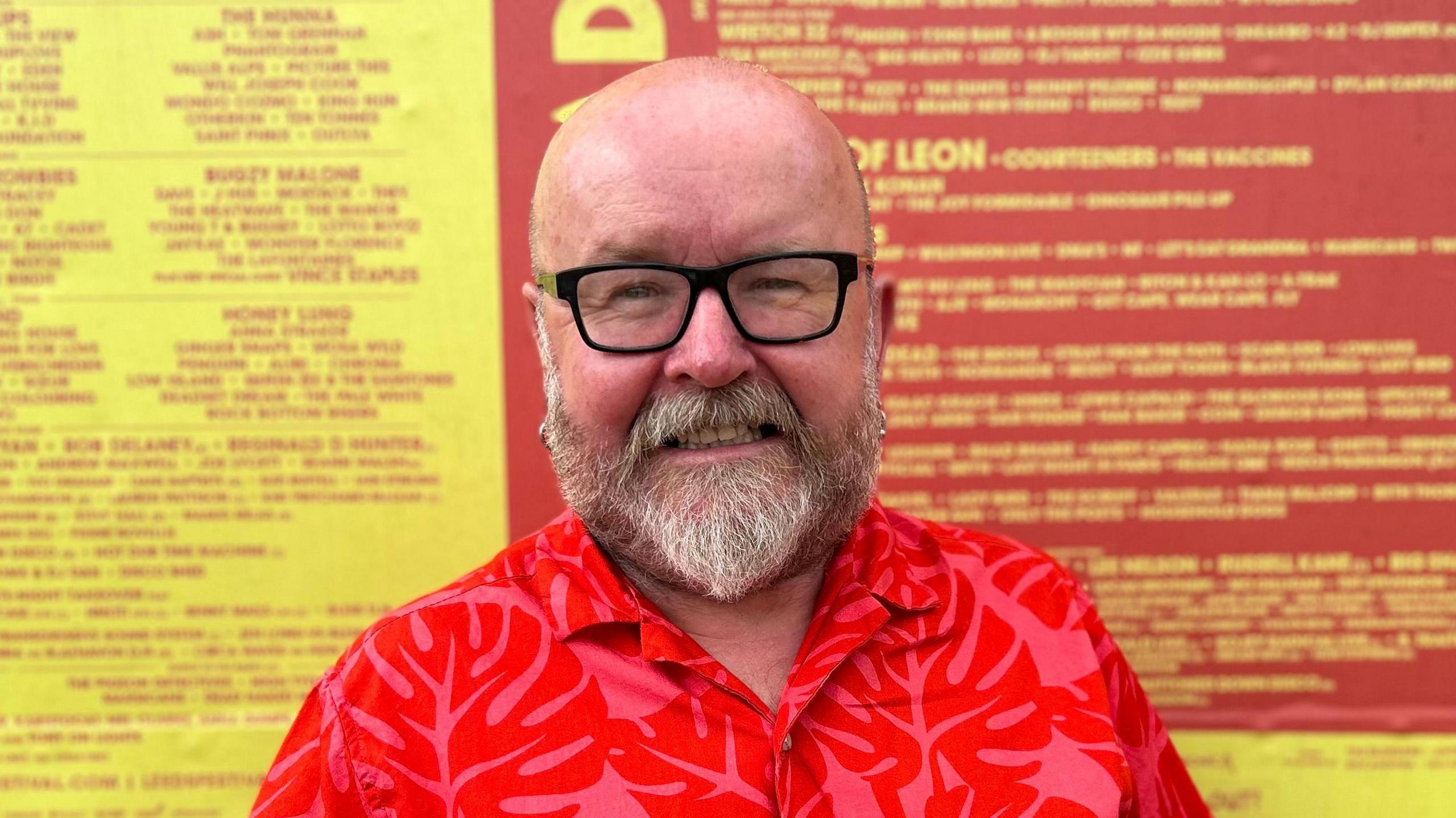 Ian Carroll is wearing a red and pink shirt. He is bald, wearing black-rimmed glasses and has a white beard.