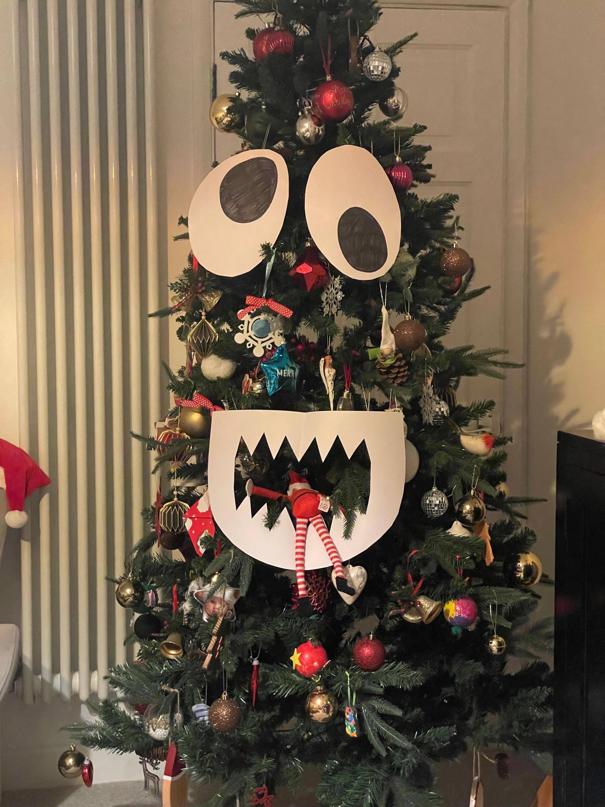 A Christmas tree has two googly eyes and a mouth. The elf is inside the mouth.