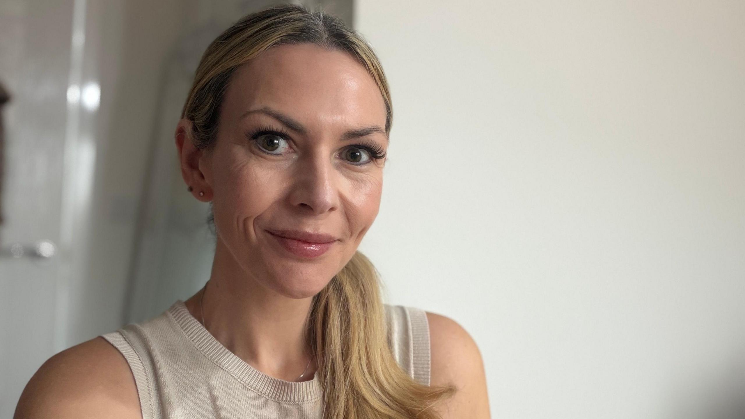A head and shoulders shot of a white woman with long blonde hair in a ponytail over her left shoulder who is smiling at the camera. She's wearing a beige vest top. 