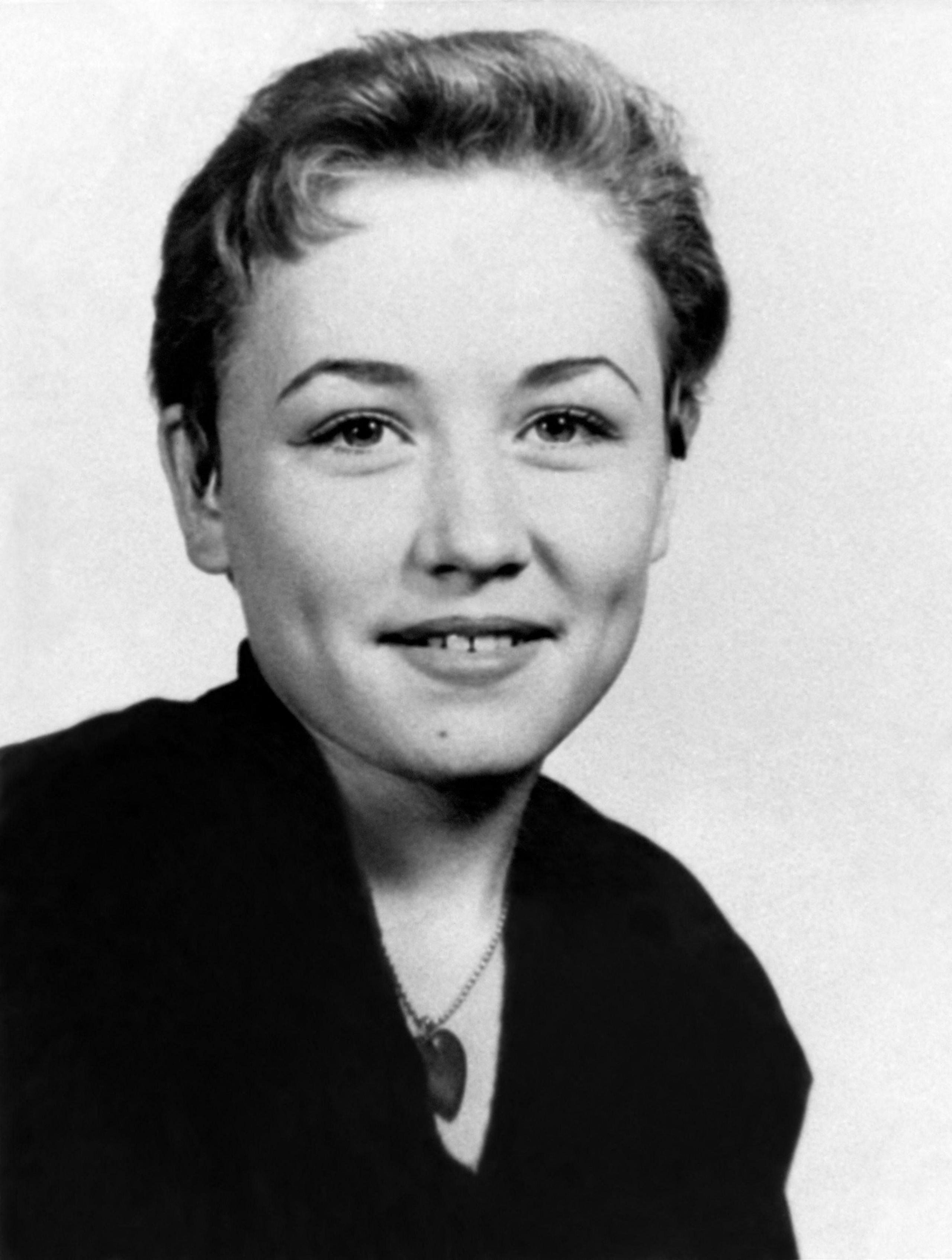 Dolly Parton, age 14, with short hair and a slight smile, in a black and white high school portrait., She wears a dark shirt and pendant necklace.