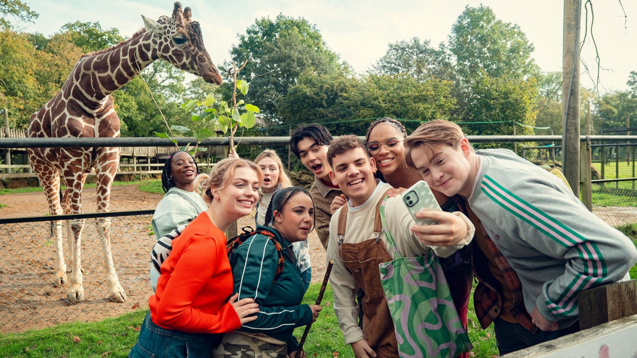 The cast of Heartstopper gather together for a selfie with Bashu the giraffe who is in his enclosure