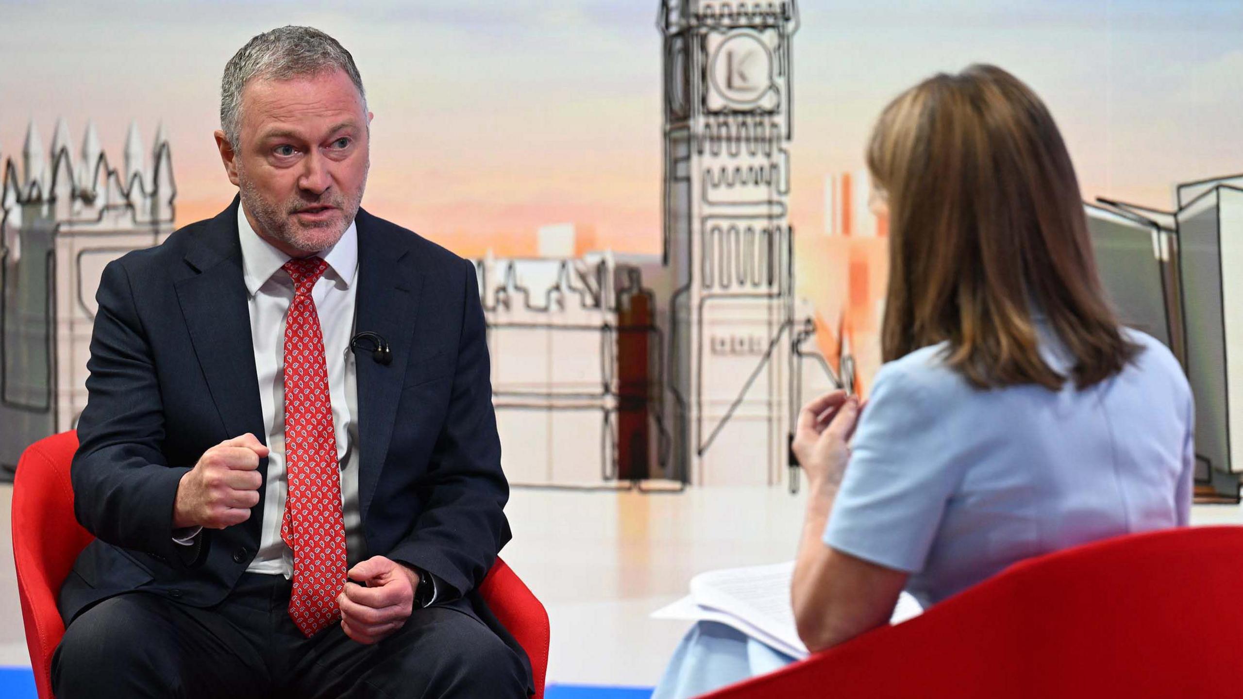 Housing, Communities and Local Government Secretary Steve Reed appearing on the Sunday with Laura Kuenssberg, with presenter Victoria Derbyshire standing in. The pair sit on red bucket chairs. Reed wears a black suit with a white shirt and a red tie. Derbyshire is wearing a pastel blue dress with short sleeves. She has shoulder length, dark blonde hair. 