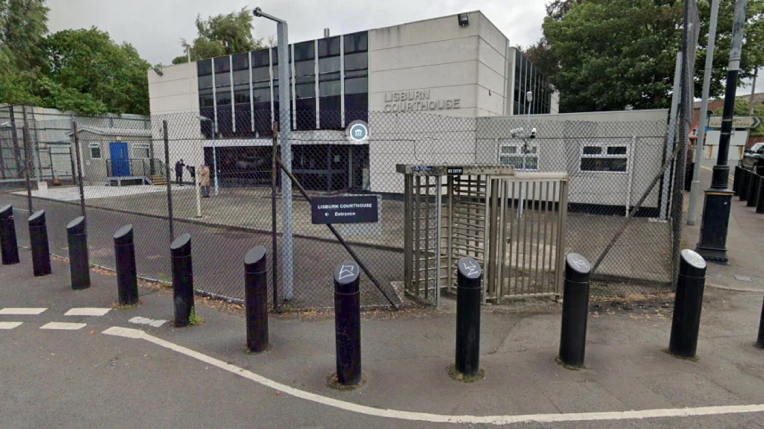 The outside of Lisburn Magistrates' Court. it is a white building with dark glass panels on the side and surrounded by wire fencing.