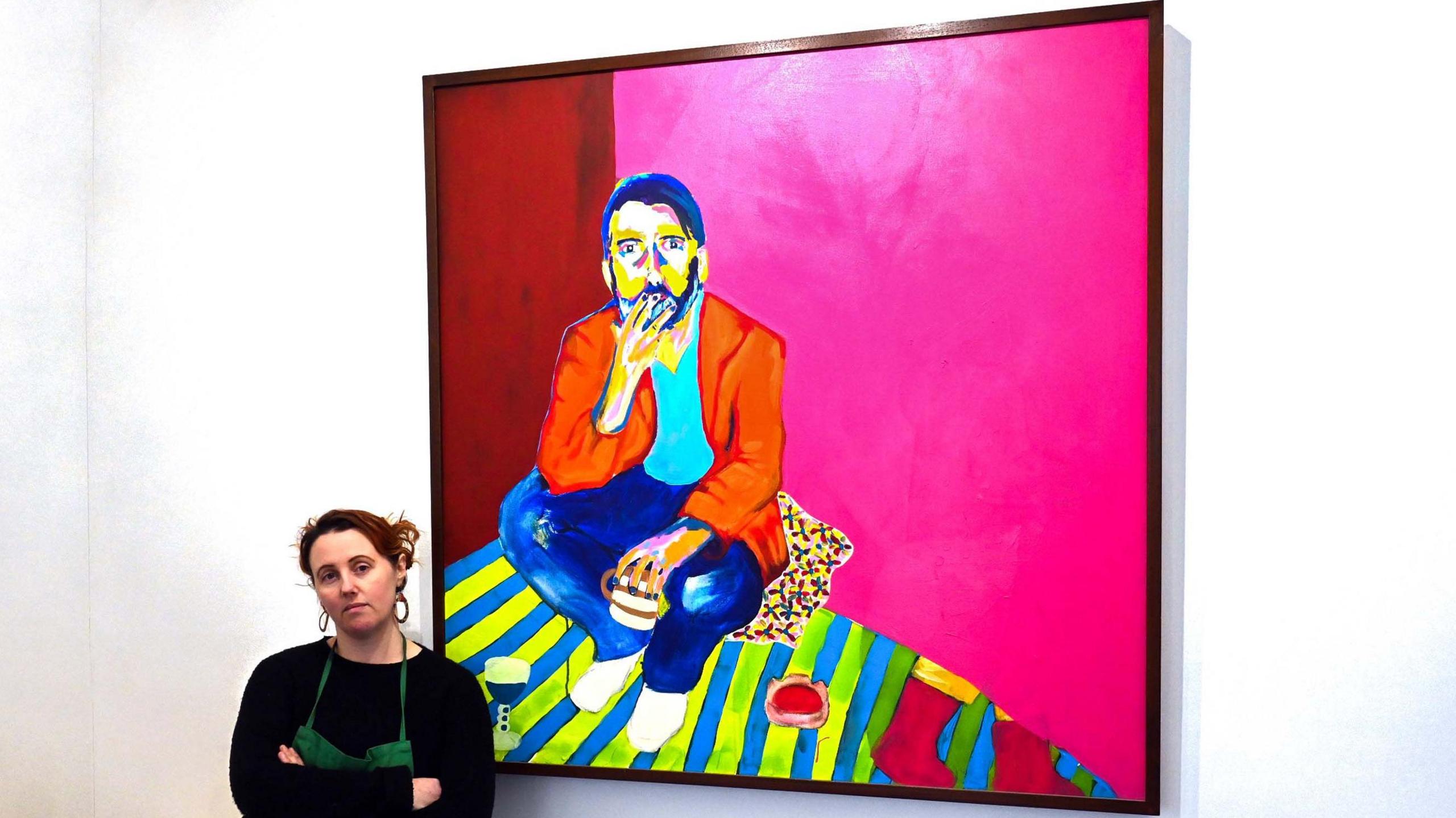 Natalie, with arms folded in front of one of her portraits, showing a bearded man sitting down, smoking and holding a mug