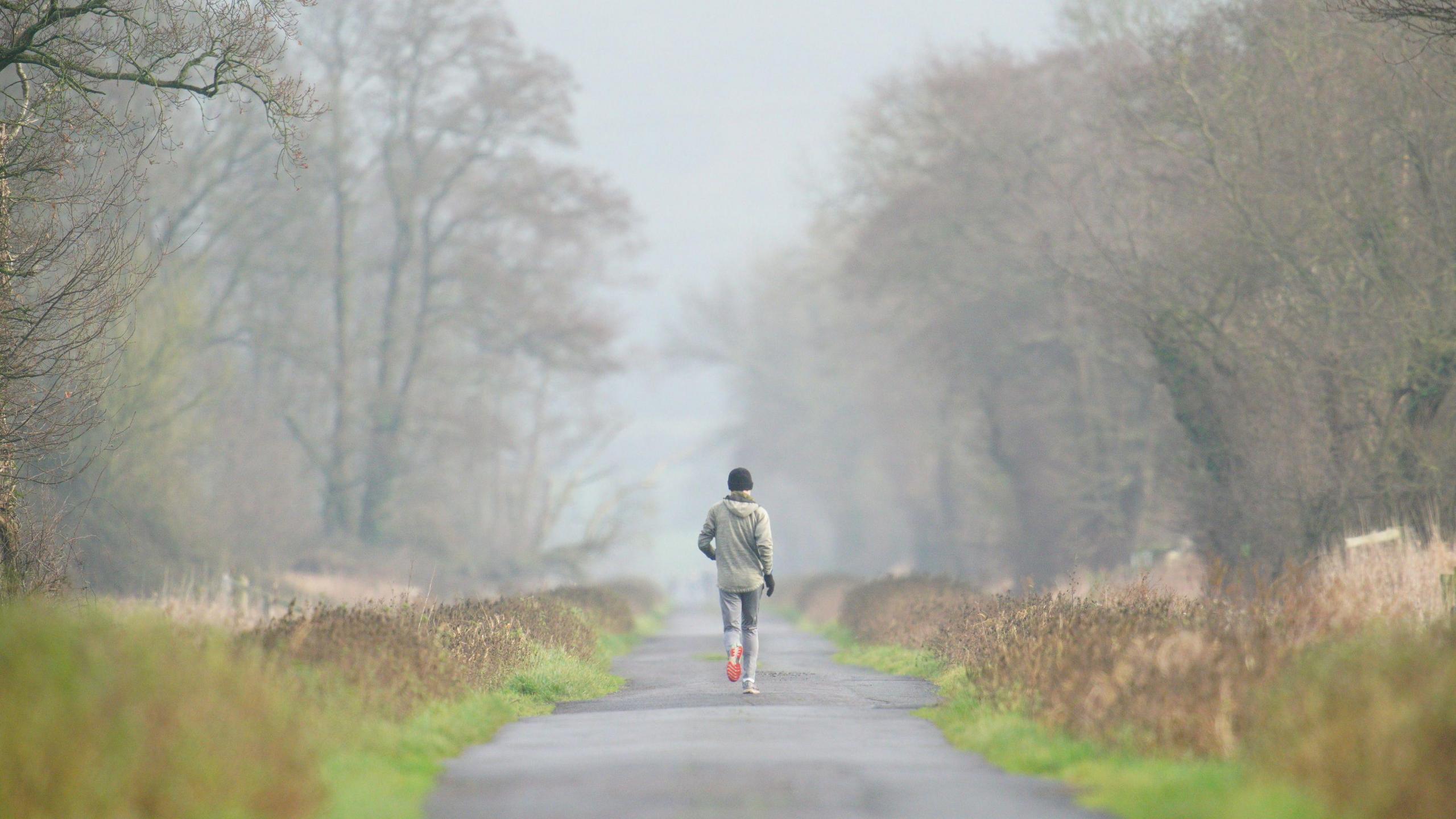 A person out jogging on a foggy morning. They are pictured with their back to the camera. They are wearing a black beanie, a beige jacket, black gloves, grey trousers and trainers with bright orange soles. They're travelling down a lane in Glastonbury which is lined by trees and grass.