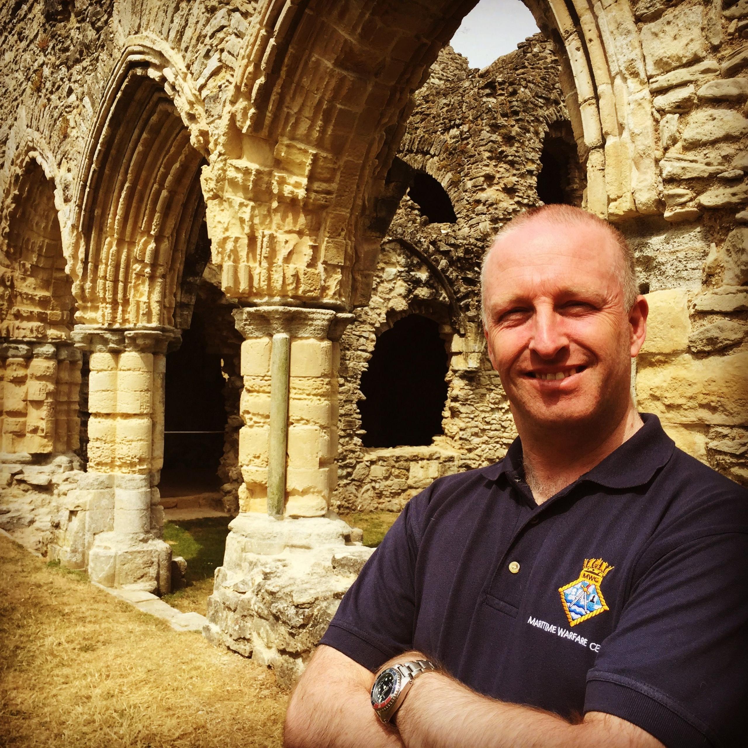 Richard Jones standing in front of historic ruins. He has his arms folded and is wearing a black polo shirt with a logo on it. He is bald and is smiling at the camera.