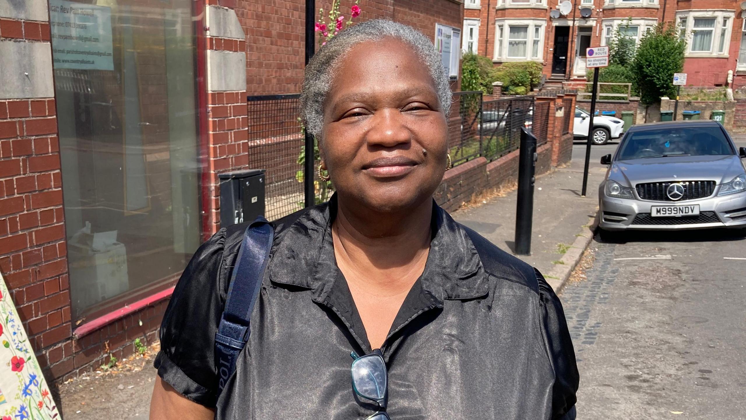 A woman in a black top stands in front of a church by a side road with a silver car parked behind her.