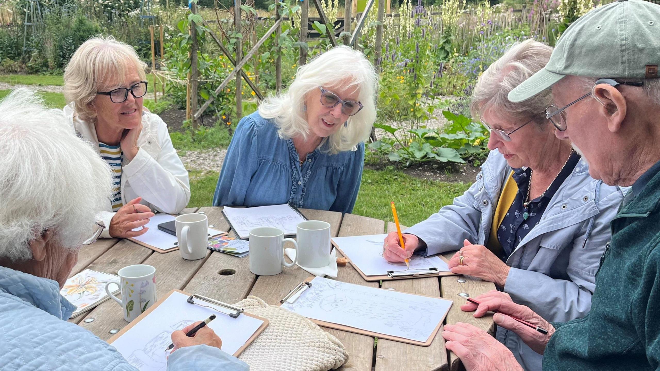 Four women and one man sitting around a round wooden table in a garden. There are plants and flowers in the background. The people all have clip boards and paper with drawings on on the table in front of them and two of the ladies are drawing with pencils, while the teacher, a woman with white shoulder length hair, glasses and a denim shirt looks on. There are also four empty mugs on the table.