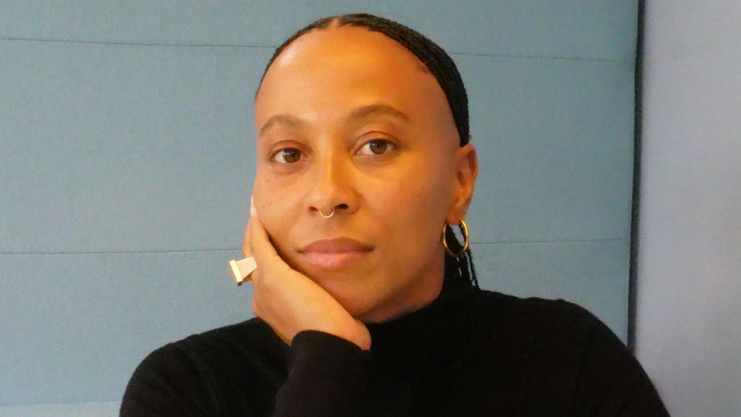 Ella Janneh looks to camera with her hand on her chin. She is wearing a black jumper and has a gold ring on her finger, a nose ring and a gold hoop earring.