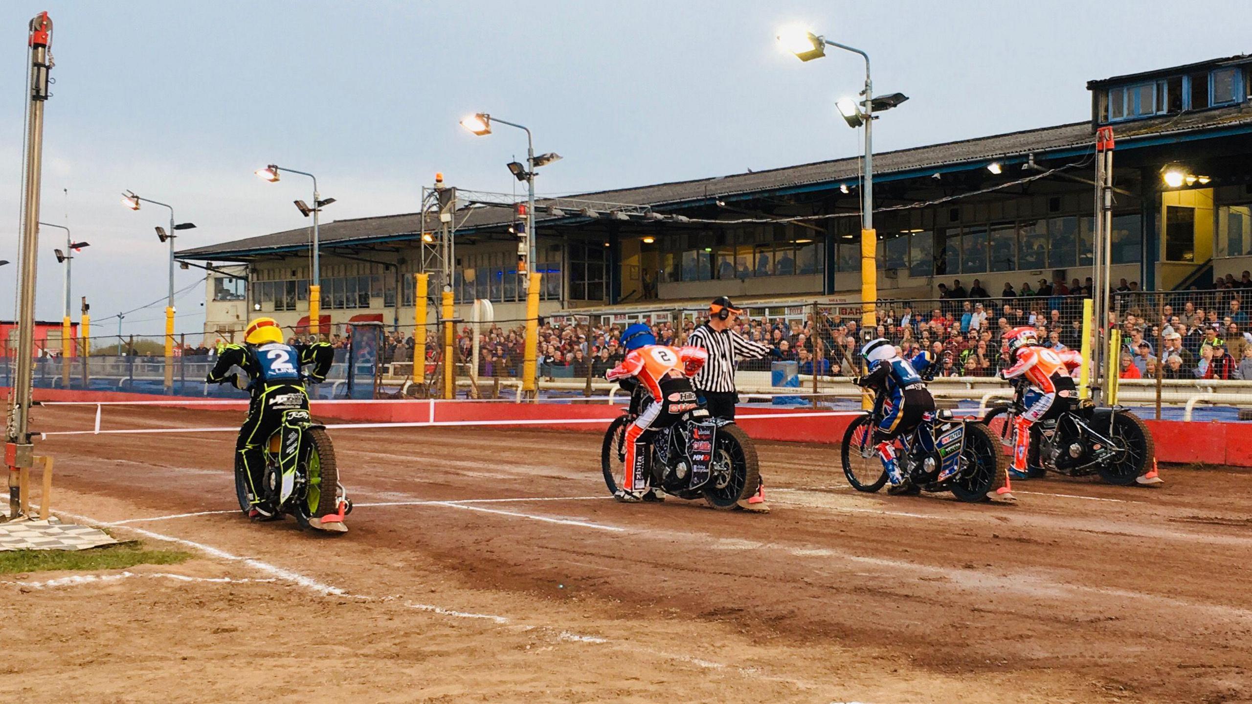 A speedway track with four riders waiting to get under way at the starting tape. Stadium lights and a grandstand full of fans in the background.