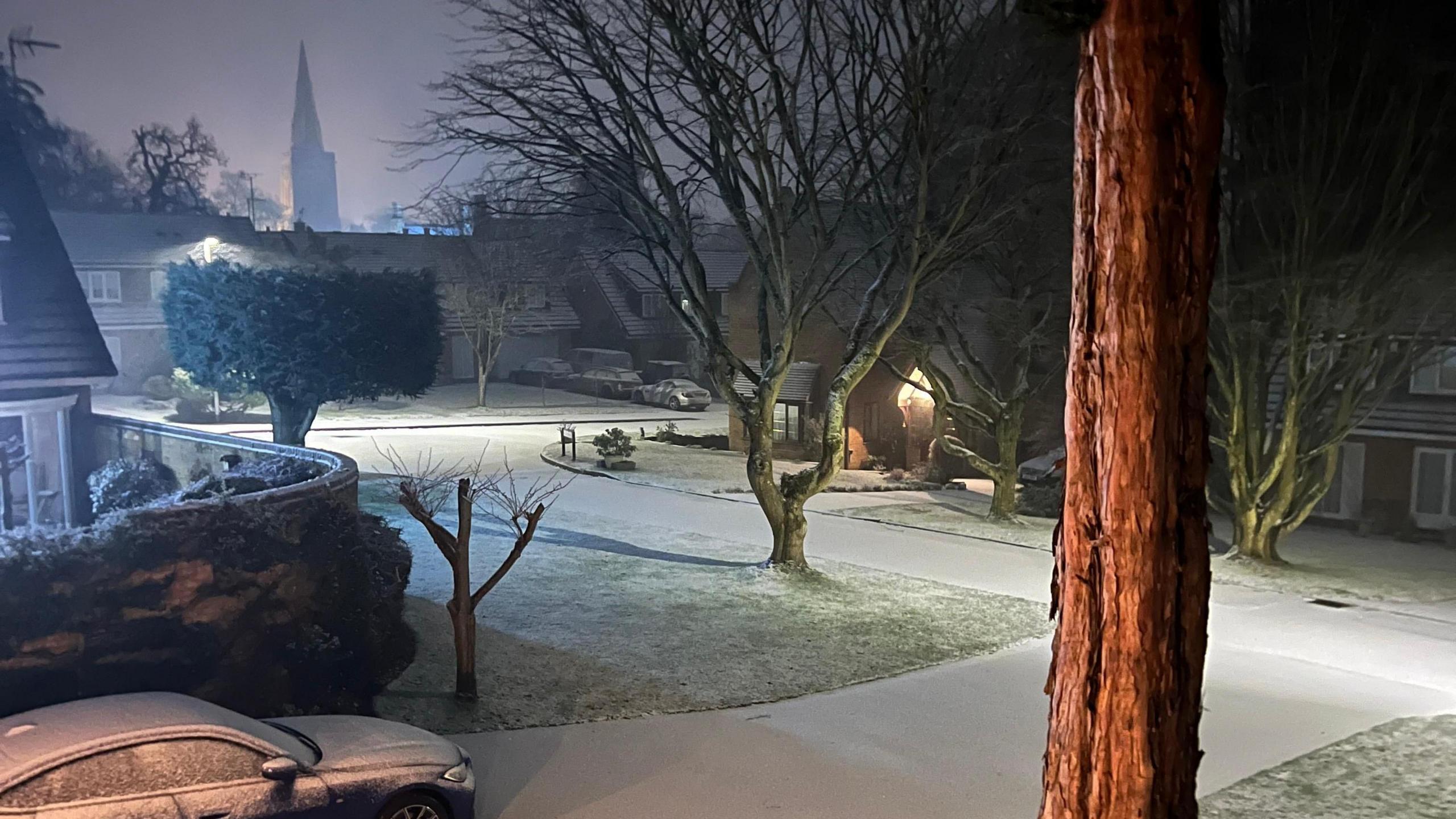 Residential roads covered in snow, a church is in the distance, a tree trunk to the right and snow on paths and on trees. 