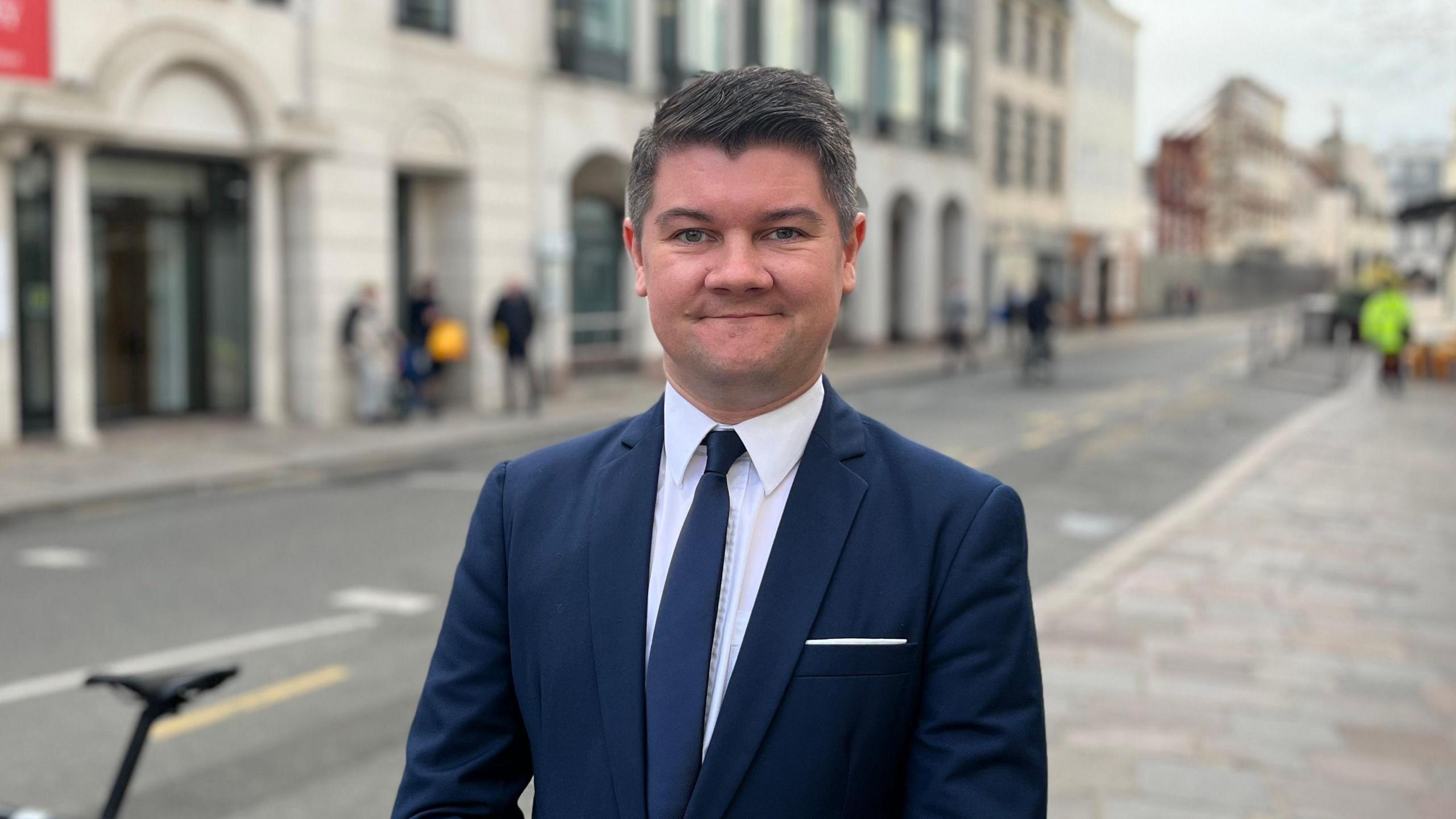 A man wearing a navy suit, navy tie and white shirt is standing in front of the  States of Jersey building smiling at the camera. Behind him but slightly blurred are a road, people on pavements and buildings.