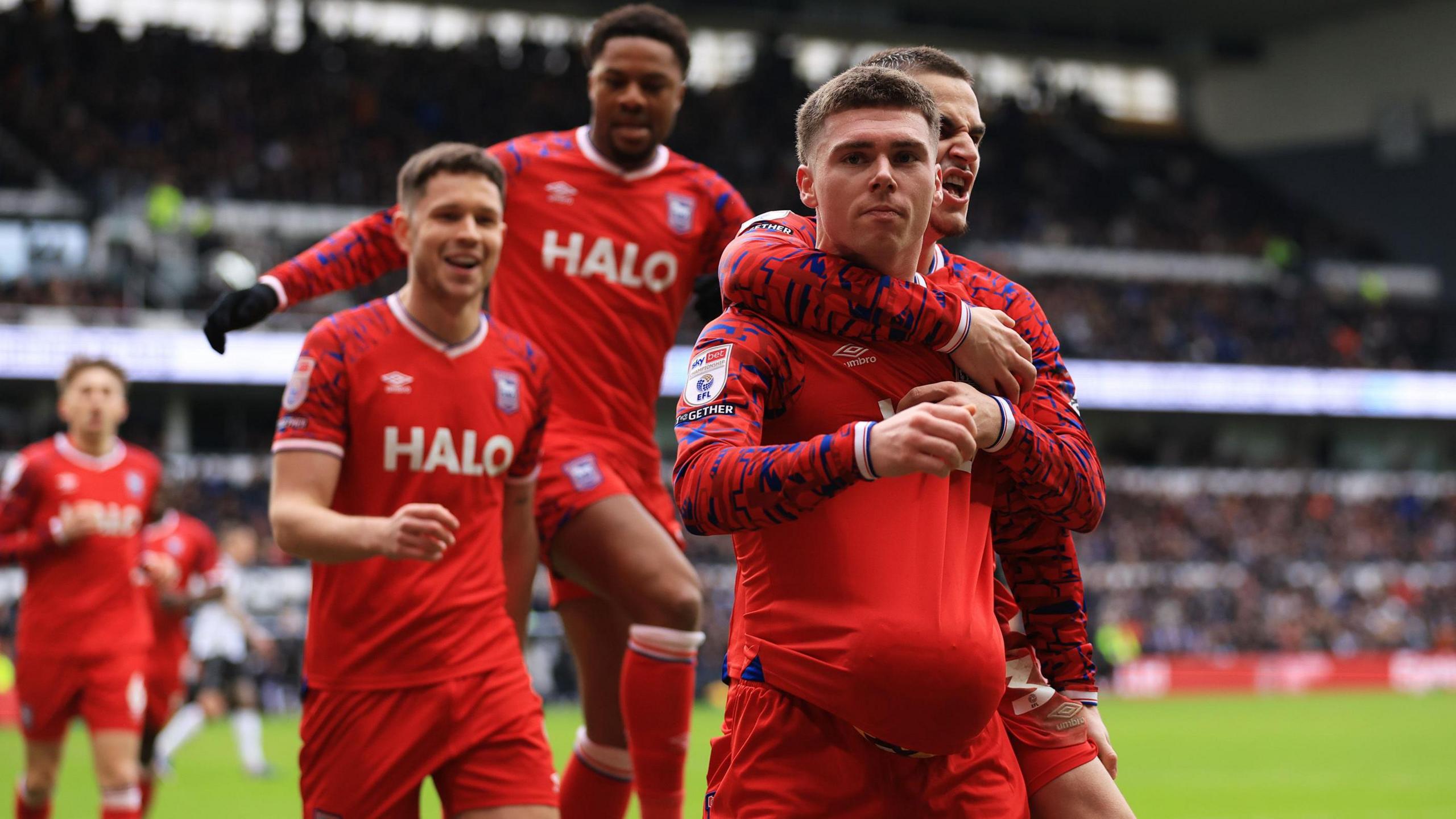 Ipswich Town players, wearing the club's red away kit, celebrate with goalscorer Leif Davis, who has a ball up the front of his shirt, at Pride Park