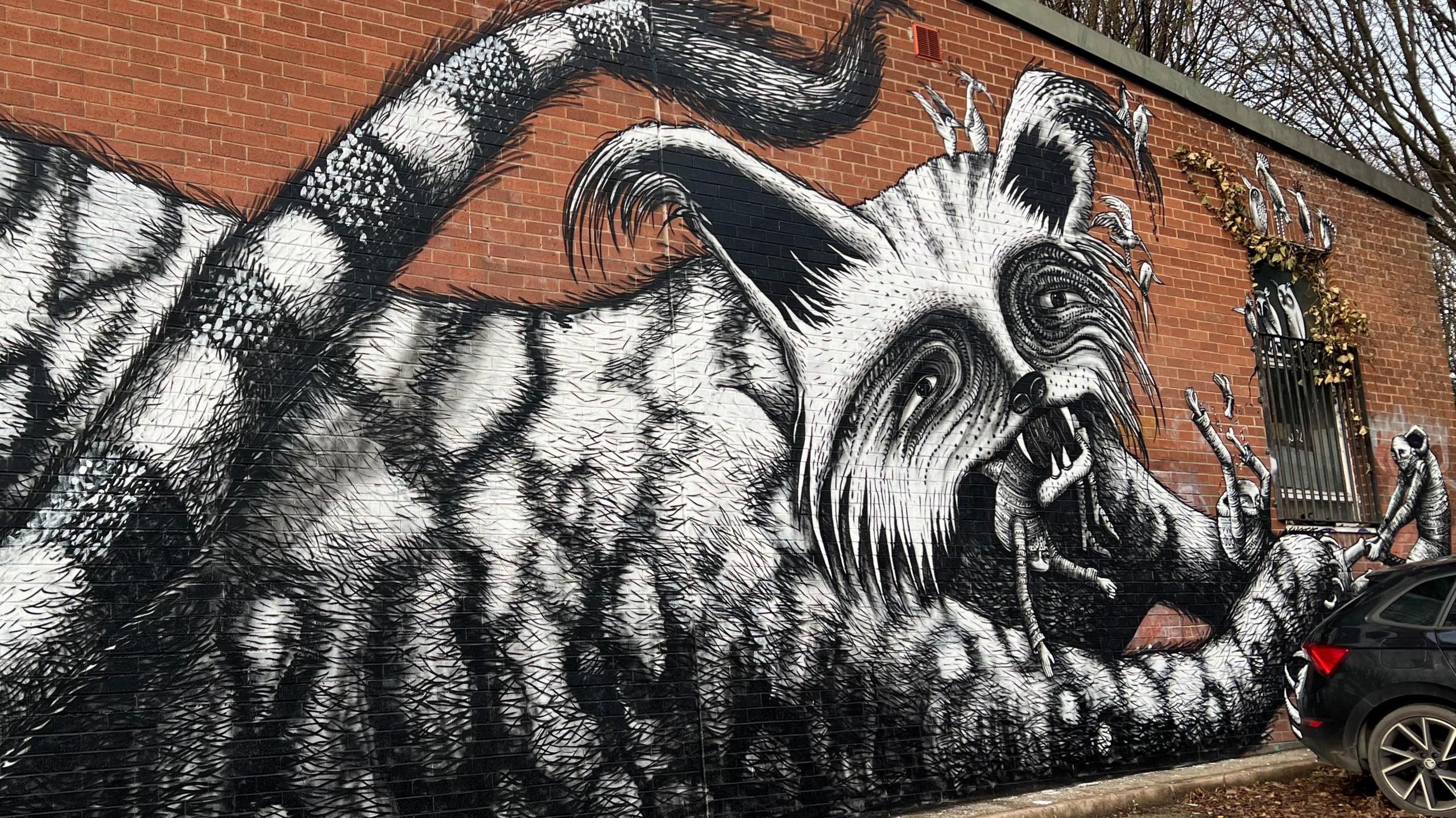 The mural, which is on a brick wall. A black and white depiction of a giant, grotesque cat who is trying to eat a group of human beings. One of the people is hanging from its mouth. Another is being attacked by its claws, whilst a third tries to pull them free.