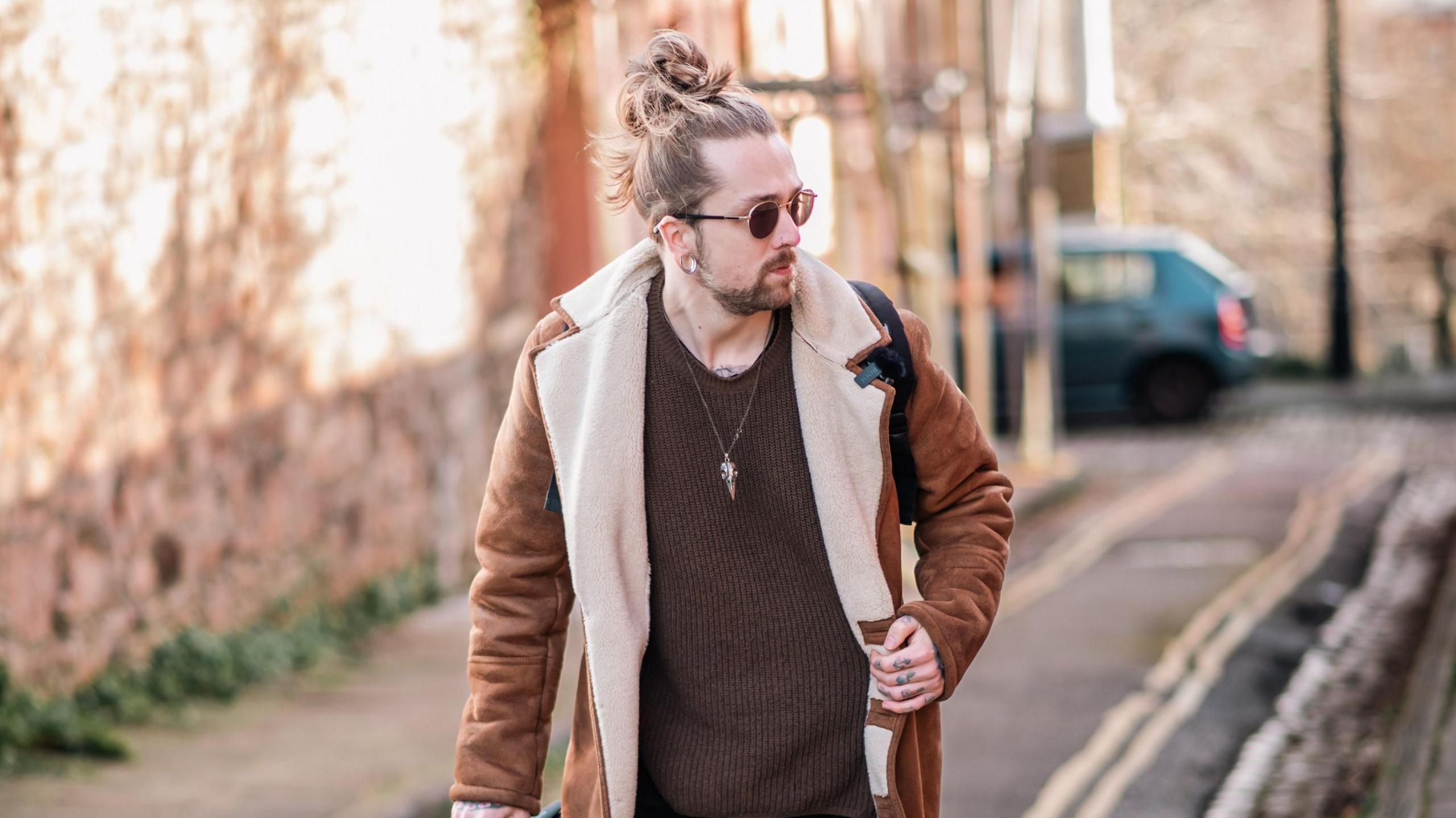 A professional picture of Mitchell Lucas. He is photographed looking away from the camera wearing sunglasses, while walking down a narrow street. 