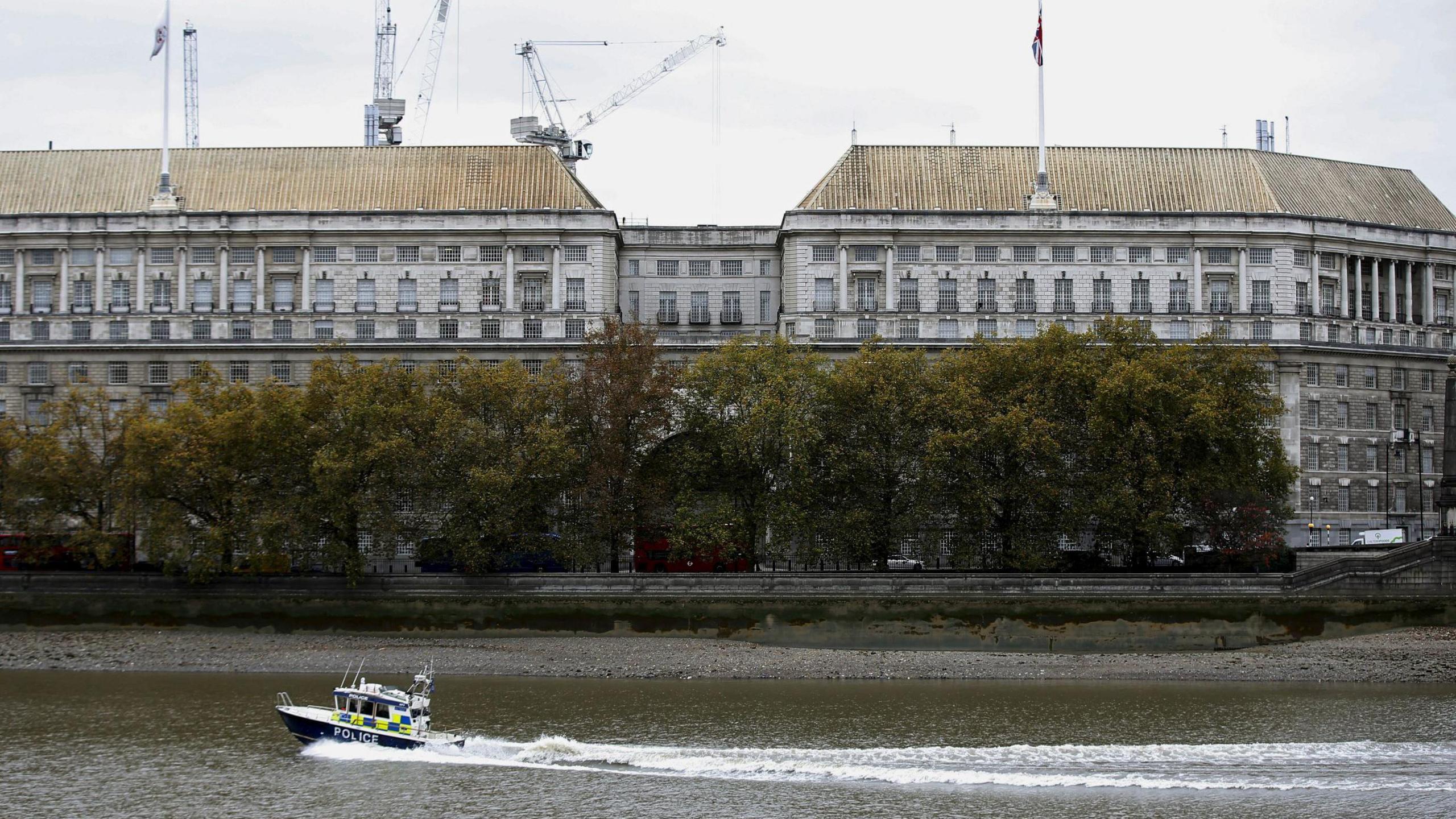 A view across the river Thames of MI5 headquarters, an imposing neo-classical building in grey stone. Flags are flying from the roof and construction cranes can be seen behind it. Part of the frontage is obscured by trees along the river, and a police boat is passing by on the water.