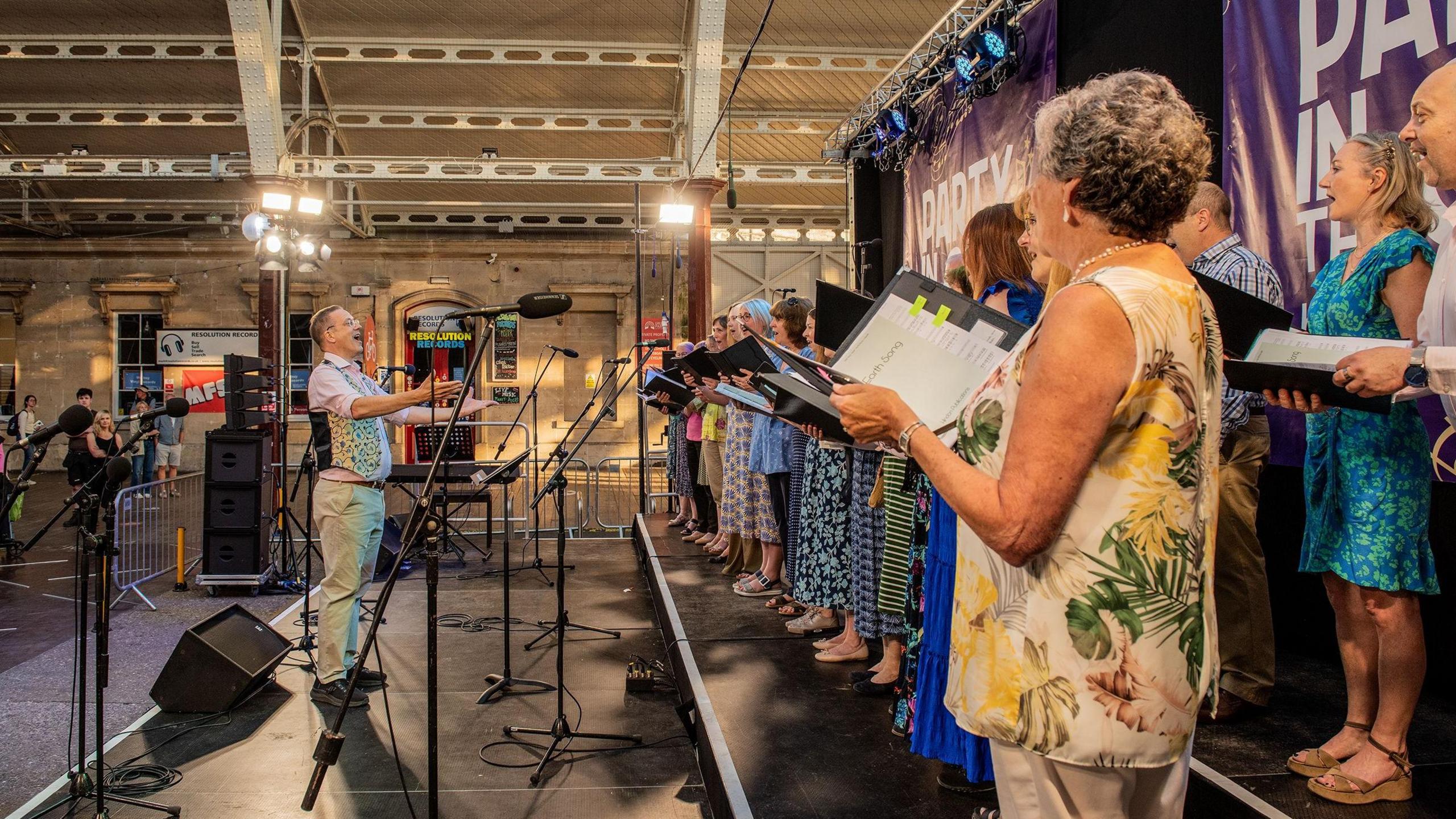 A male choir leader stood in front of a choir. The choir are standing opposite him holding folders of sheet music. There are several microphones in front of the choir. Nobody is looking at the camera - the choir leader is looking at the choir and the choir is looking at him.
