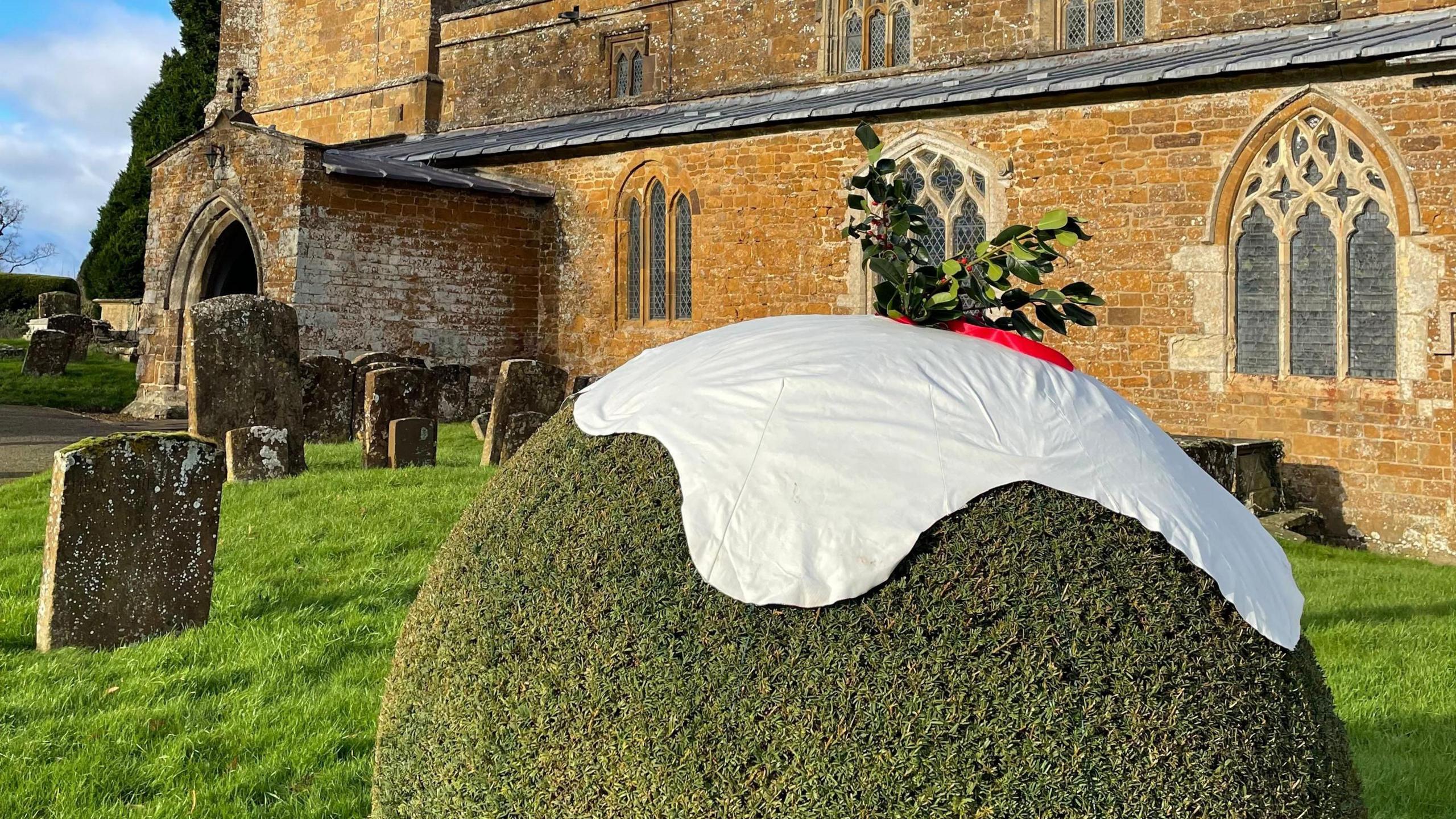 A large circular green bush sits in front of a church. A white sheet has been put on top of it to look like the sauce on top of a Christmas pudding. Some green foliage with red berries has also been placed on top.