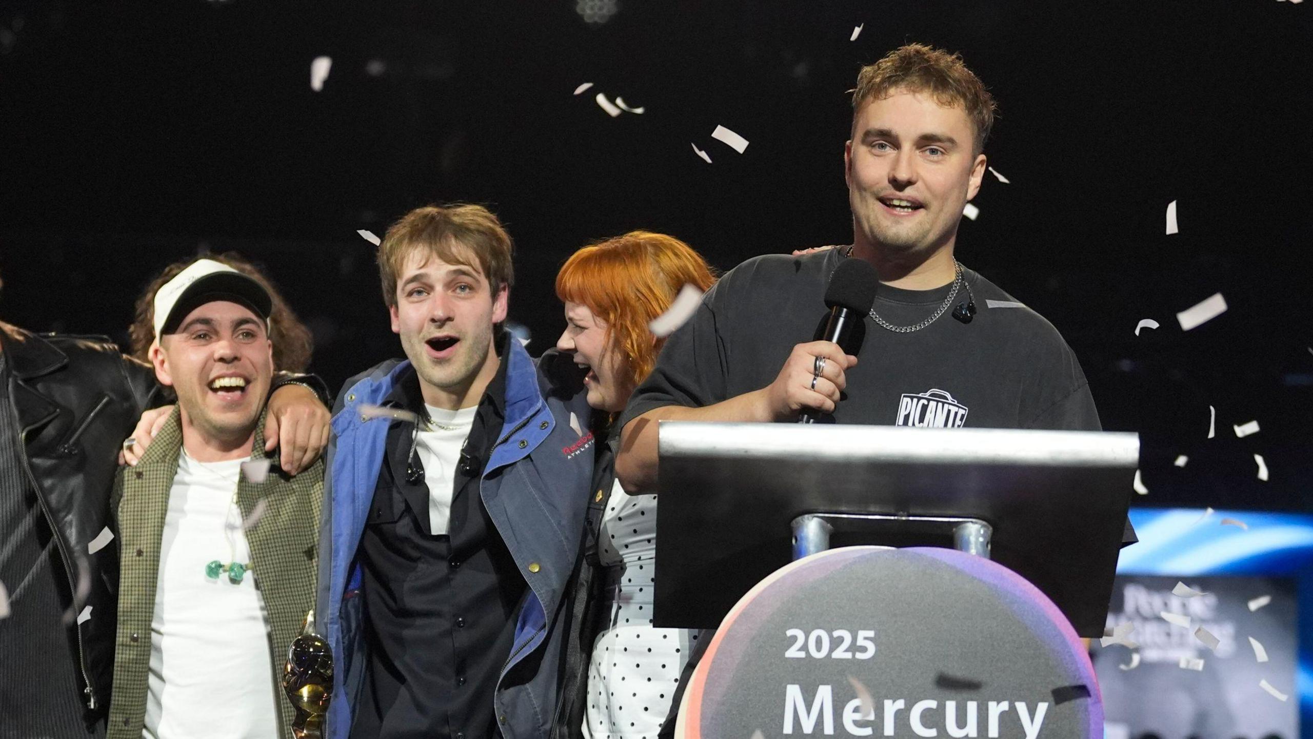 Sam Fender at the podium after winning the Mercury Prize in Newcastle. He is holding a microphone and smiling. Confetti is raining down around him and his band, who are standing behind him with their arms around each other.