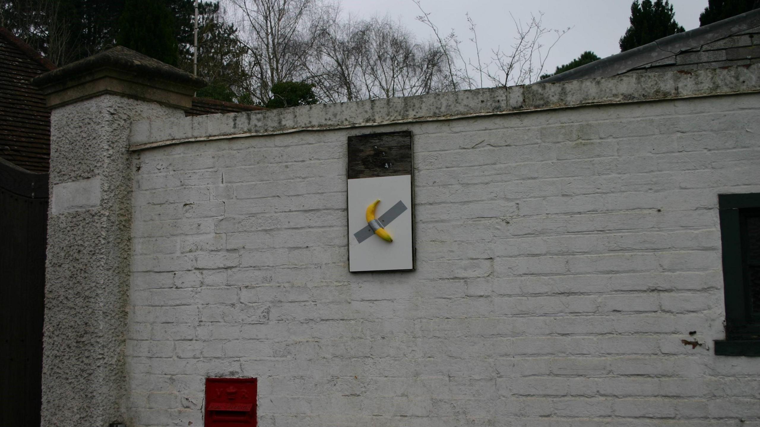 A banana duct-taped to a square white board is affixed to a white brick wall