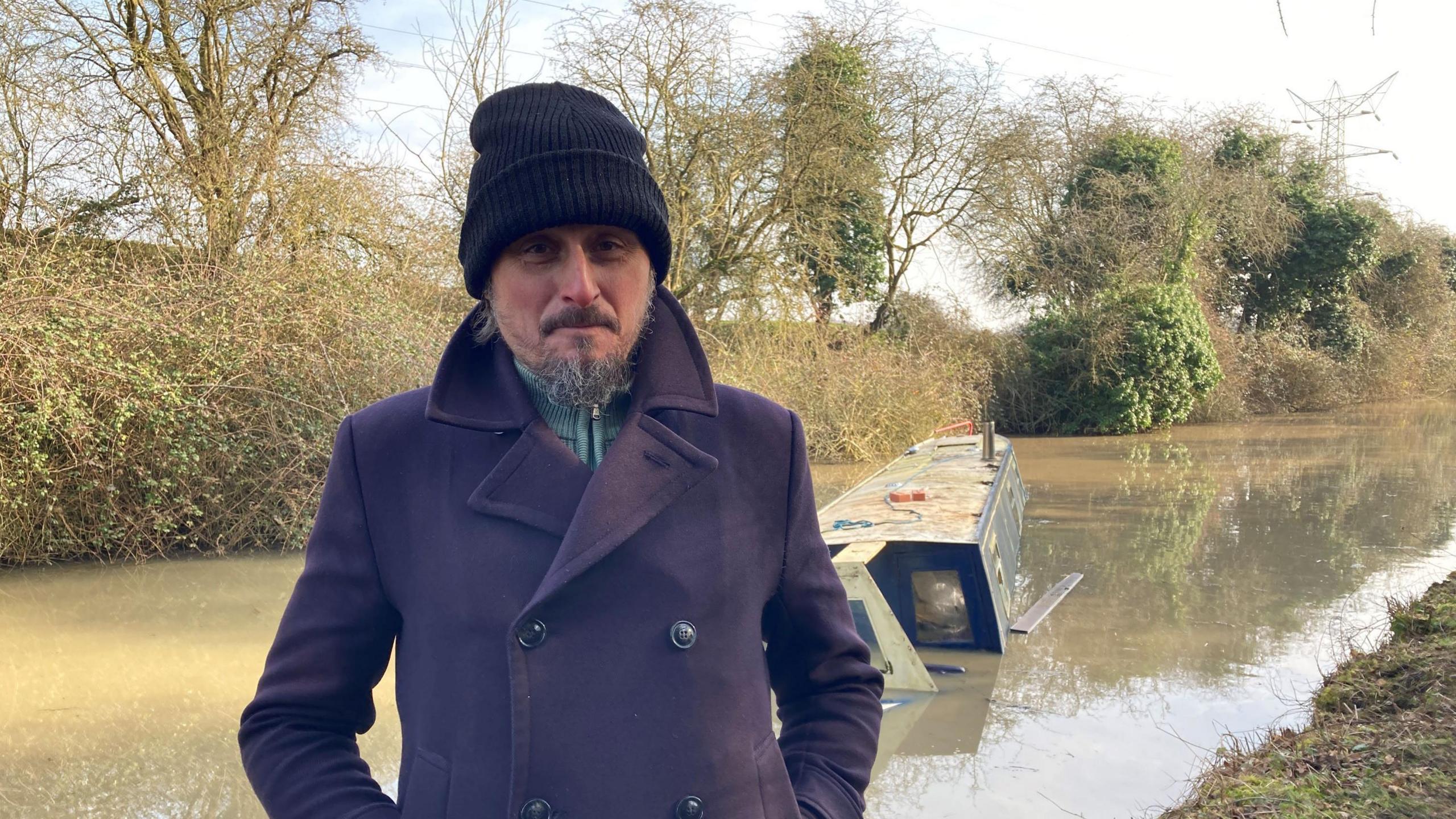 A man in a hat and pea coat stands by a canal in front of a sunken narrowboat with his hands in his pockets. 