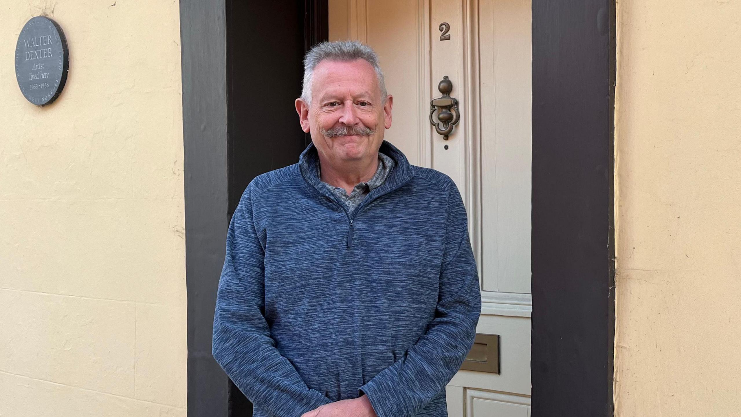 Kim Leonard is standing in front of a grey door with the number 2 written on it. He has grey hair and a moustache and is wearing a blue jumper.