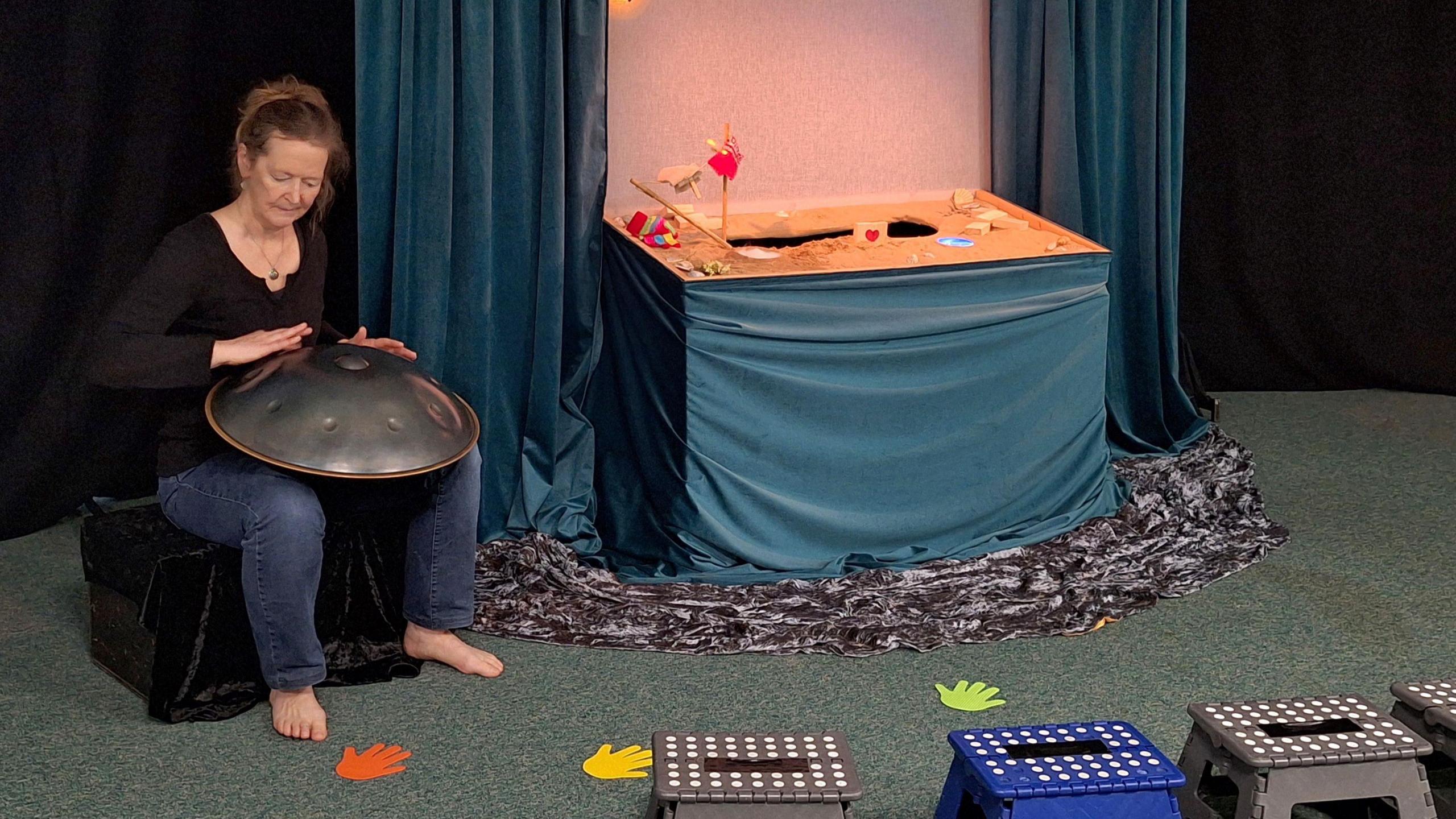 Lempen Puppet Theatre performs at Skipton Puppet Festival. A woman sits on a stool, with bare feet, blue jeans and a black top. She holds a steel drum, and sits next to a small puppet stage.