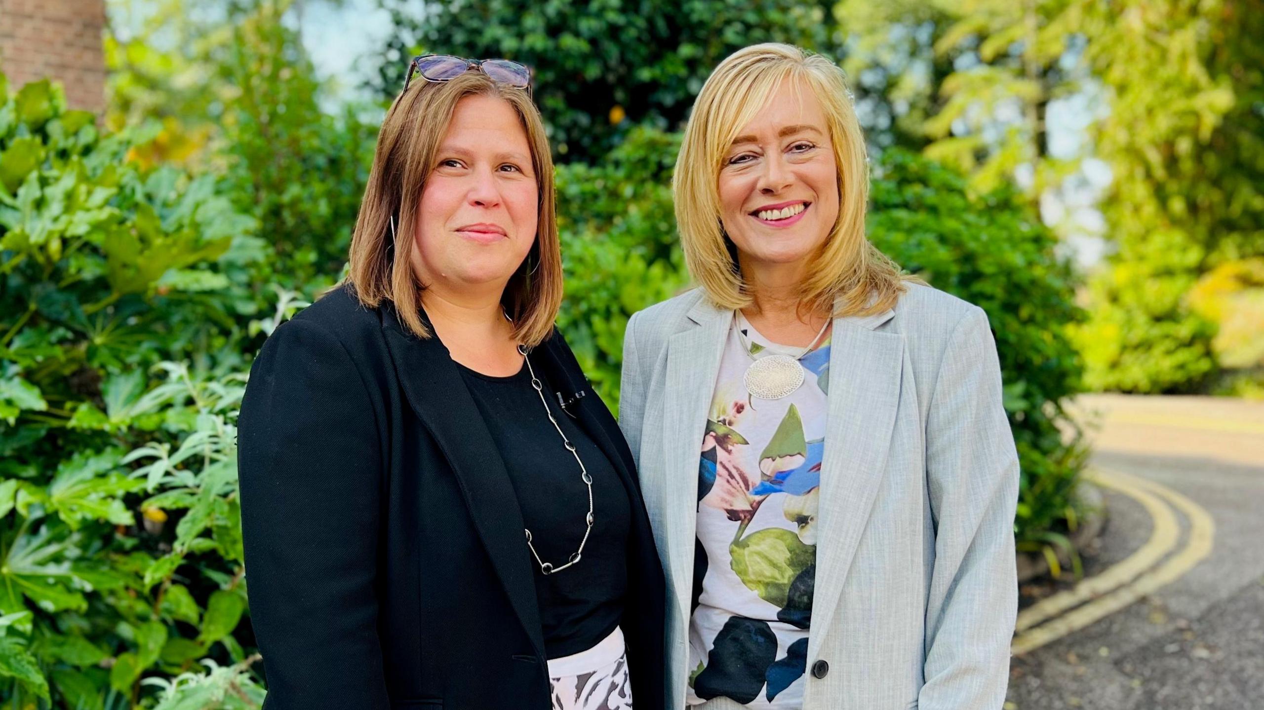 Standing outside in front of greenery in the sunshine are the two female Vice Chancellors who have negotiated the merger. On the left is Professor Georgina Randsley de Moura, the interim Vice Chancellor of the University of Kent wearing a black jacket and top. On the right is Professor Jane Harrington the Vice Chancellor of the University of Greenwich wearing a pale grey jacket with a patterned floral top. Both women are smiling